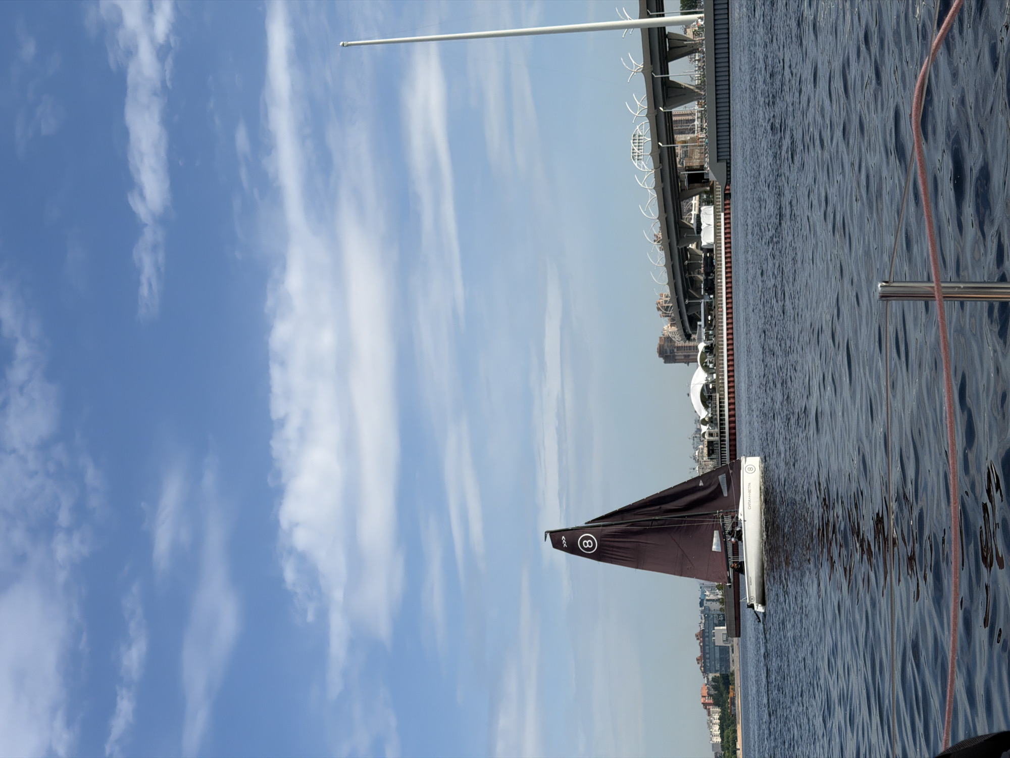 A white sailboat with a dark brown sail, marked with the number "8" in a white circle, sails on dark blue water under a partly cloudy sky. In the background, a modern elevated highway with distinctive curved white supports spans across the horizon, separating the water from a cityscape featuring buildings and a tall white pole. The scene appears to be on the Neva River in Saint Petersburg, Russia. Two individuals are visible in the sailboat. The foreground shows a railing with a pink rope.