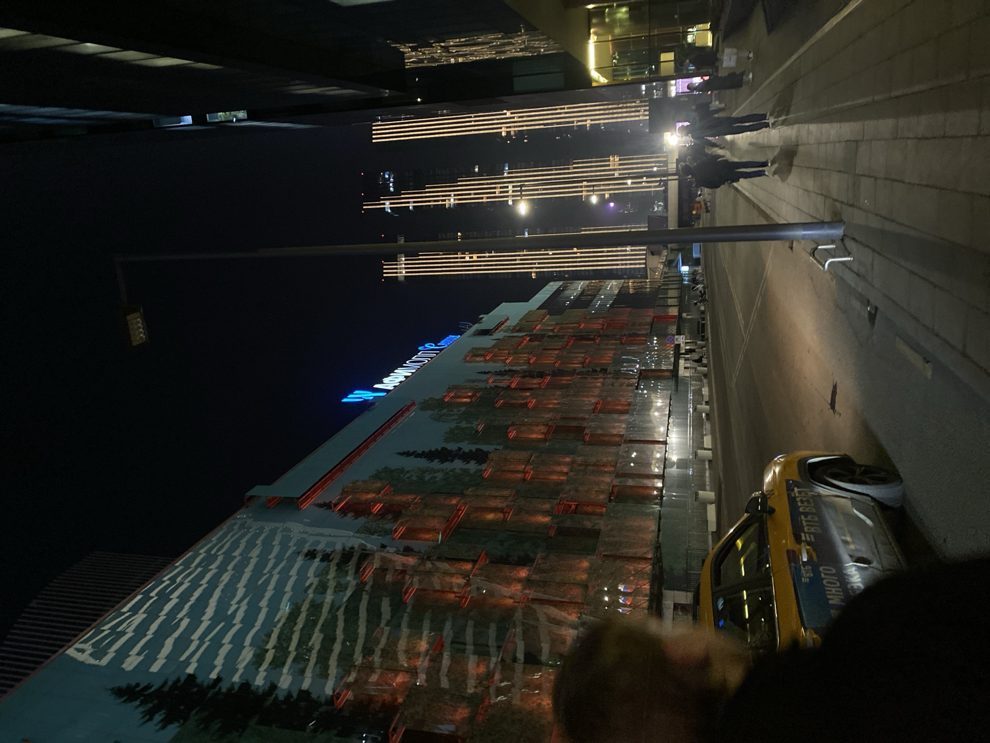 The image depicts a night scene in Khimki, Russia, featuring the Aquilon Resort hotel. The hotel's facade is illuminated with lights and a decorative pattern resembling trees. A yellow taxi is parked on the street beside the hotel. Pedestrians are visible walking along the sidewalk. Street lights provide additional illumination. A bridge or overpass is visible in the background, along with other buildings and lights.