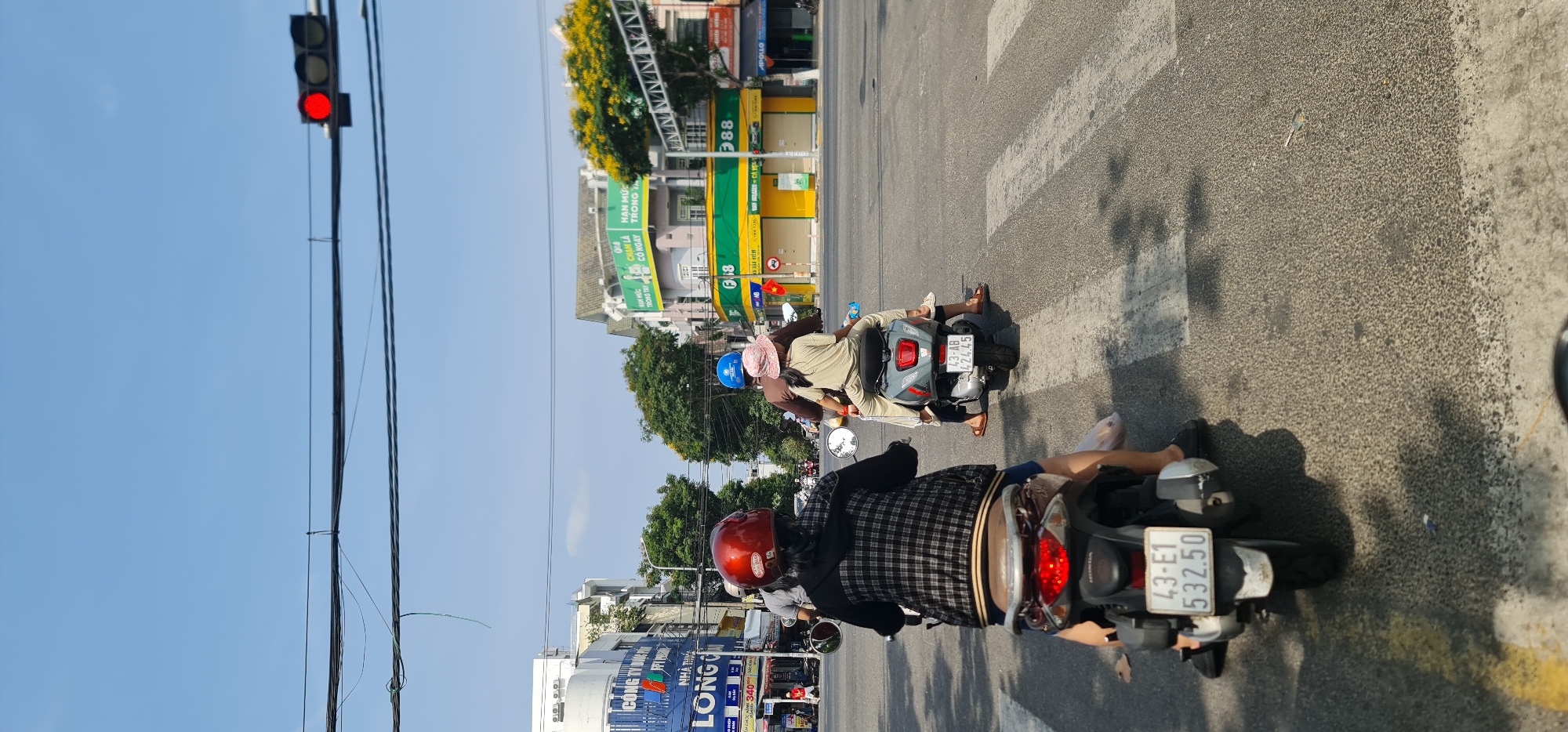 This image captures a bright, sunny street scene in Đà Nẵng, Vietnam, at what appears to be an intersection, with vehicles stopped at a red traffic light. The perspective is from behind a person on a motorcycle, looking forward.
In the foreground, the back of a brown Honda scooter is visible, with its red taillight illuminated and license plate displaying "43-E1 532.50." The rider is wearing a red helmet and a black and white plaid shirt. Ahead of this scooter is another gray scooter, with two individuals. The person in the back is wearing a light-colored outfit and a pinkish hat, while the driver wears a blue helmet. Its license plate reads "43-AB 424.45." White zebra crossing lines are painted on the asphalt road, and shadows from overhead structures and possibly trees are cast across the ground.
Beyond the stopped motorcycles, the street is lined with commercial buildings. A prominent green and yellow building with the large number "88" is visible, along with a small Vietnamese flag flying nearby. Another building further down has a blue sign with partially visible Vietnamese text, including "CÔNG TY" and "LONG." Overhead, numerous power and communication lines crisscross against a clear, light blue sky. A red traffic light is clearly illuminated, indicating vehicles are halted.
Visible text on storefronts includes "APOLLO" on a dark building, and fragments like "HAN MỨC," "TRONG TÂY," and "CÓ NGAY" on the green and yellow building. The license plates "43-E1 532.50" and "43-AB 424.45" clearly reference Đà Nẵng. The scene depicts a typical busy daytime street in an urban Vietnamese setting.