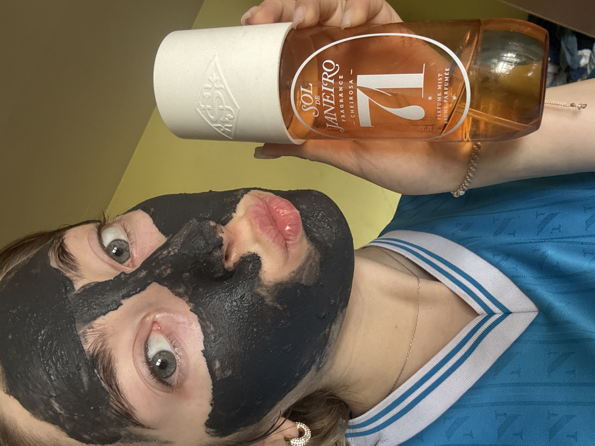 A person with a black facial mask poses with a bottle of Sol de Janeiro '71 fragrance mist. The person is wearing a blue shirt with white stripes around the collar. They are also wearing a necklace and an earring. The bottle of fragrance mist is held in the person's hand. The background is a blurred yellow wall. The city and state are unknown.