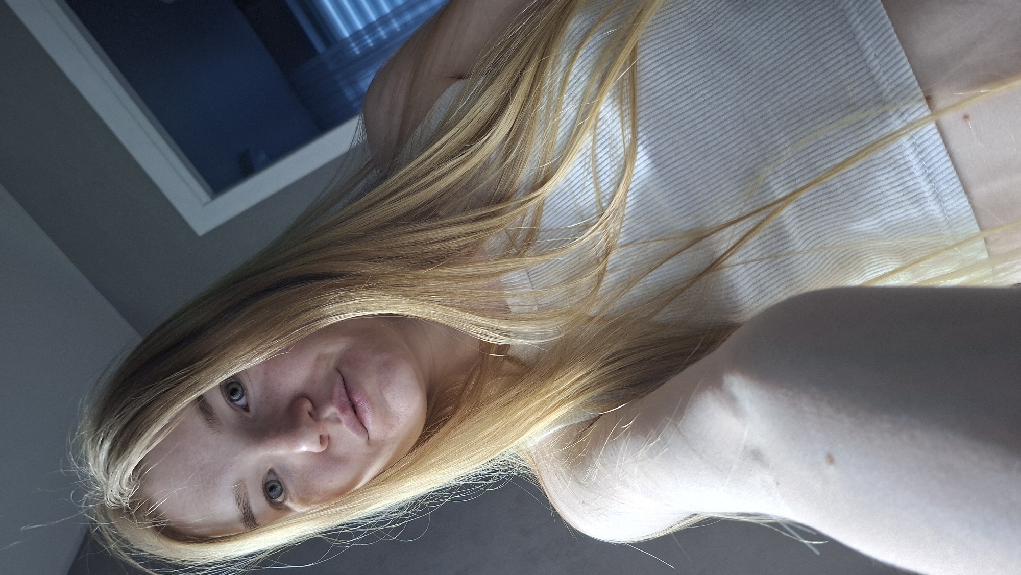 A young, light-skinned person with long blonde hair, wearing a white ribbed tank top, is photographed from a low angle. The person's face is visible, and they appear to be looking at the camera. The background is partially obscured, but a window with blinds is visible. The location cannot be determined based on the image.