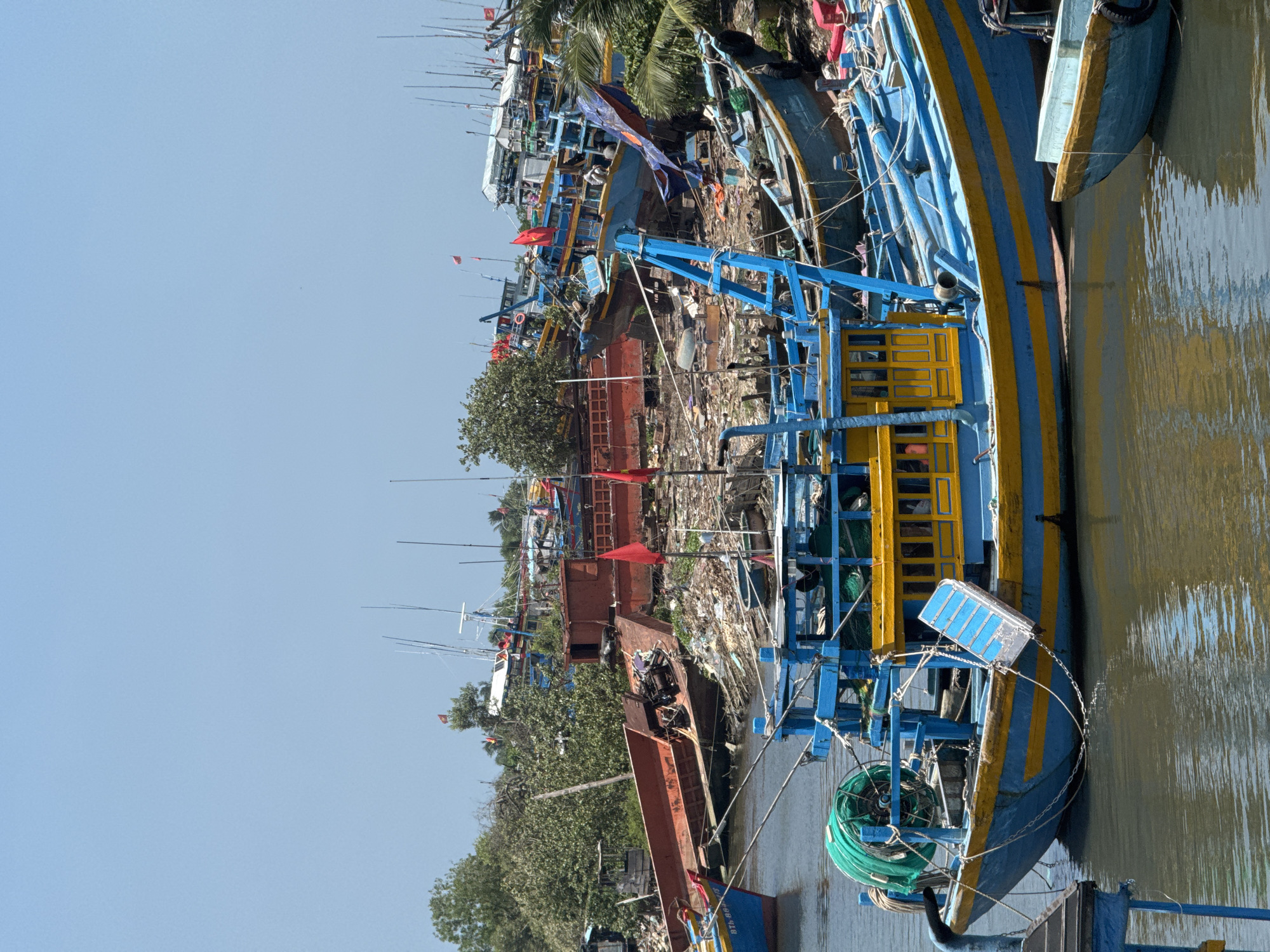 This image captures a chaotic scene in the port of Phú Thủy, Vietnam, where numerous fishing boats are moored. The foreground is dominated by a vibrant blue and yellow fishing boat with a green fishing net reel attached to its stern. To the right of this boat, a smaller, lighter blue boat with a yellow trim floats in the water.
The background reveals a dense collection of fishing vessels, many of which appear to be in disrepair or abandoned. There are several large, rusty red boats interspersed with the blue ones. The shore behind the boats is heavily cluttered with debris, including plastic waste, discarded materials, and vegetation, creating an unkempt environment. Red flags are visible on some of the boats and poles, possibly indicating nationality or affiliation. The sky is clear and blue, suggesting a sunny day. The overall impression is one of a working port grappling with the challenges of aging infrastructure and waste management.