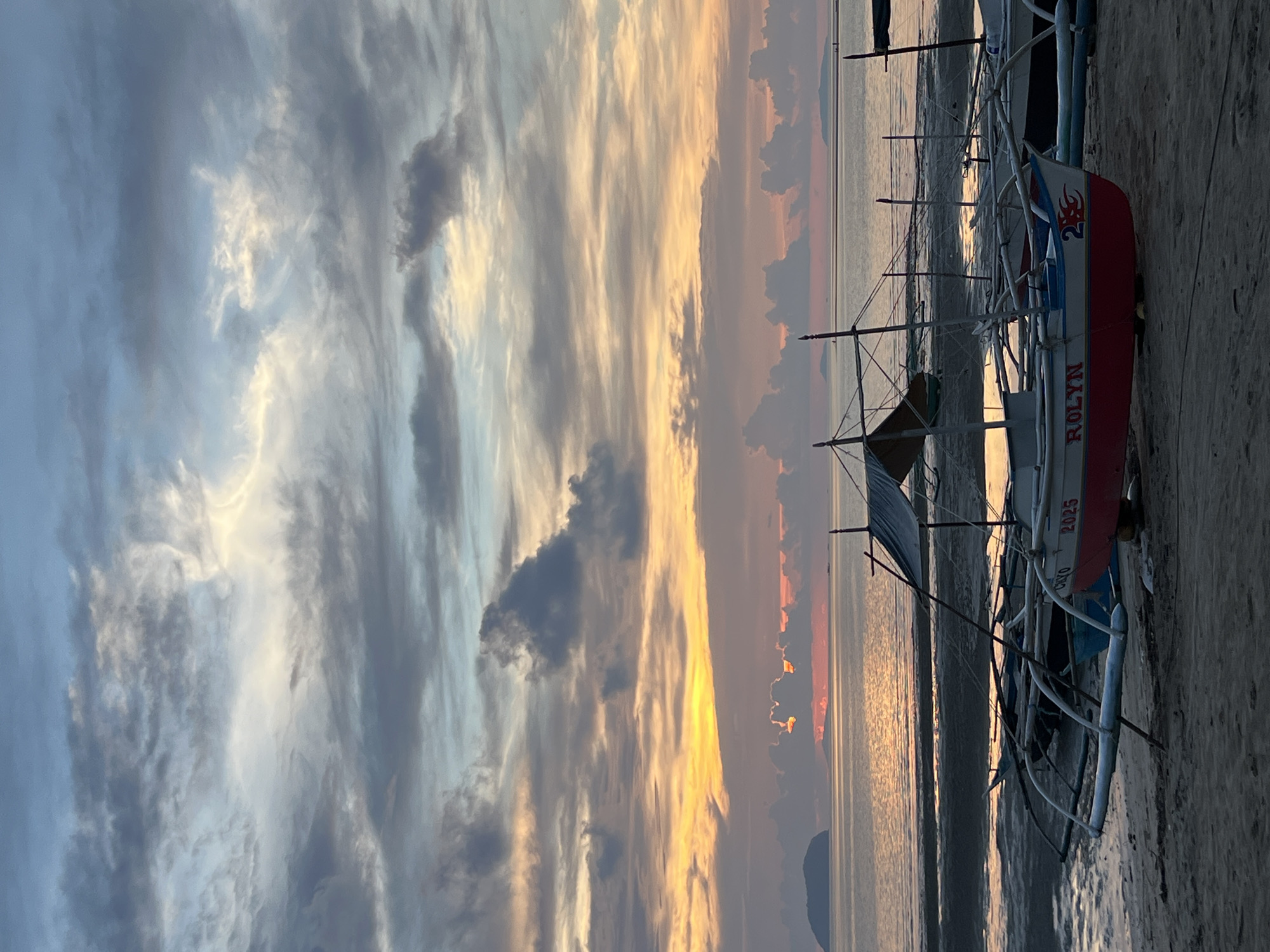 This is a vertical, eye-level, medium shot of several outrigger boats pulled up onto a sandy beach at dawn in Cuyo, Philippines. The sky is a dramatic display of clouds painted with hues of orange, pink, yellow, and blue, indicating the rising sun. The sun itself is a bright orange glow just above the horizon, peeking through a break in the clouds. The ocean is calm and reflects the colors of the sky. In the distance, a small island or landmass is visible on the left. The boats are traditional Filipino outriggers with bamboo outriggers and canvas coverings. One boat in the foreground is painted red and white with "ROLYN" and the number "2025" in red lettering. Another boat behind it is partially visible and appears to be dark blue. The beach is composed of sand and what looks like mudflats exposed by the low tide. There are no people visible in the image. The overall atmosphere is serene and peaceful, capturing the beauty of a tropical sunrise.