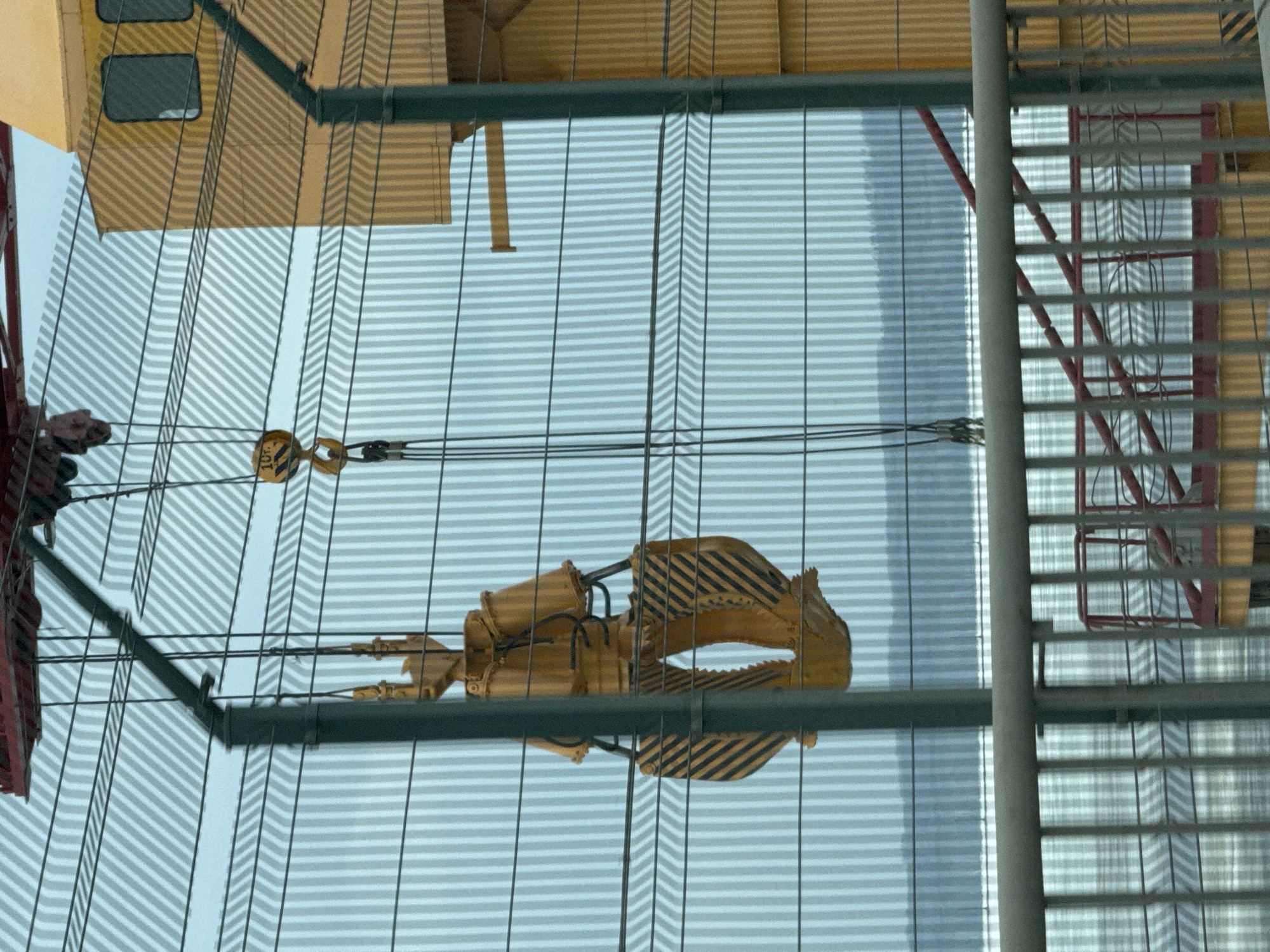 A low-angle, slightly rotated view of a construction site in Chaikovsky, Russia. The foreground features a window with horizontal blinds, reflecting a yellow and black striped mechanical grab hanging from a construction crane via multiple thin cables and a pulley. The crane's boom and machinery are visible to the left. To the right of the crane, a silver ladder with red rungs is visible through the blinds, partially obscured. The backdrop shows a cityscape obscured by the blinds.