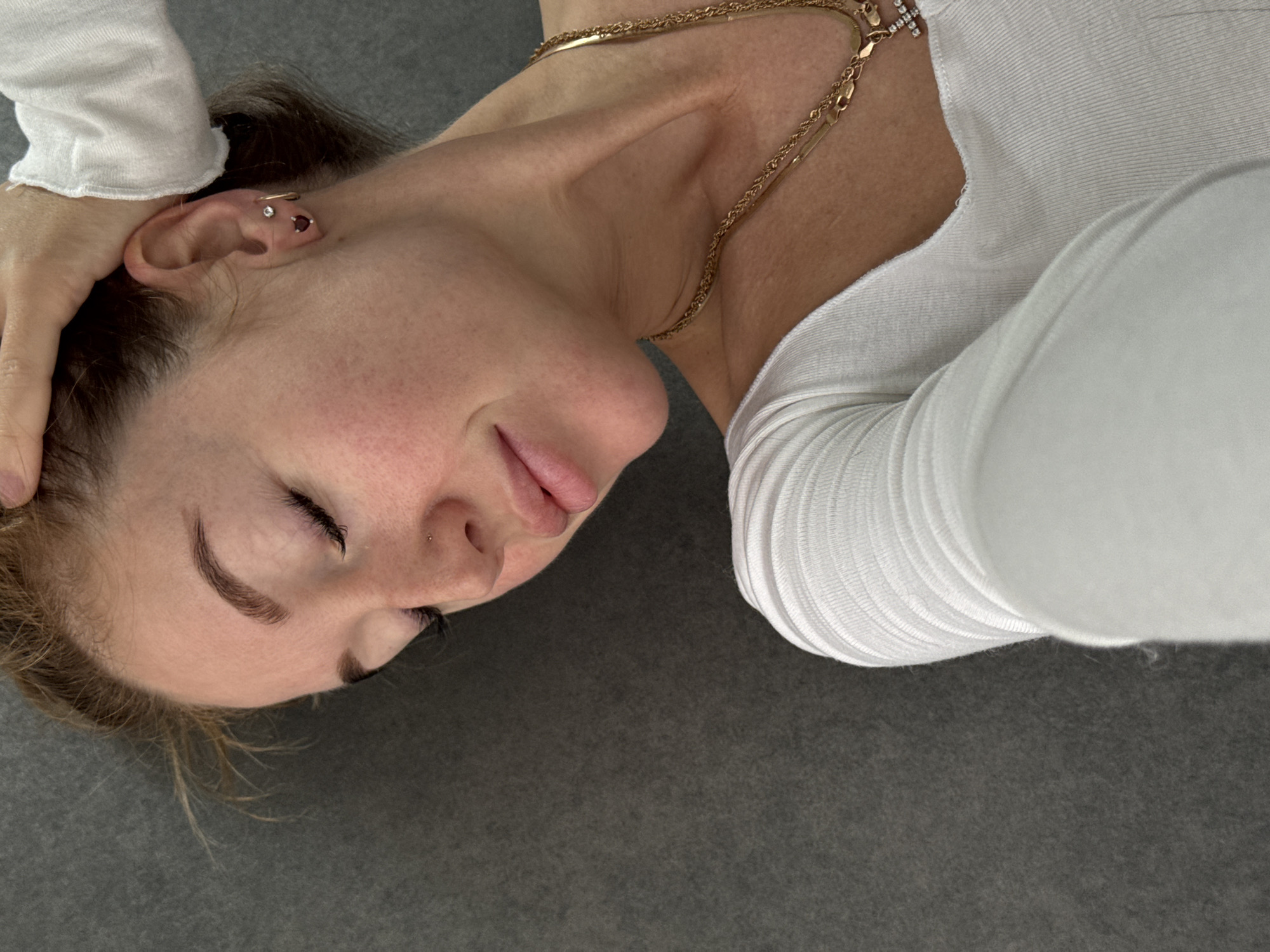 A woman with fair skin and closed eyes is lying on a gray surface. She is wearing a white long-sleeved top and a gold necklace. Her left hand is behind her head. She has multiple piercings in her left ear and a nose piercing on the left side of her nose. The location of the photo is undetermined, therefore its location in the city of Chaikovsky, Russia, cannot be confirmed.