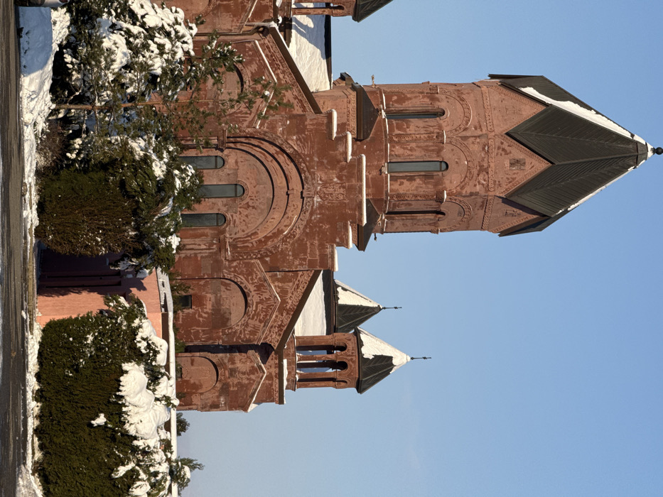 This image displays a large, reddish-brown stone church located in Rassvet, Russia, under a clear blue sky during winter. The primary structure is dominated by a central octagonal tower with a dark, pointed, pyramidal roof exhibiting significant snow accumulation on its upper facets. This tower features multiple tall, narrow arched windows. The main body of the church below the central tower is constructed from the same reddish stone, characterized by large, decorative rounded arches above sections containing smaller arched windows. To the right, two smaller auxiliary towers, also topped with dark, conical roofs partially covered in snow, are visible. The ground in the foreground is uniformly covered in white snow. Numerous evergreen trees and bushes, heavily laden with snow, occupy the lower left and mid-ground. A section of a light pink wall or building is partially visible behind the snow-covered vegetation on the right side of the frame. The scene is brightly illuminated by natural light. No individuals or active subjects are present.