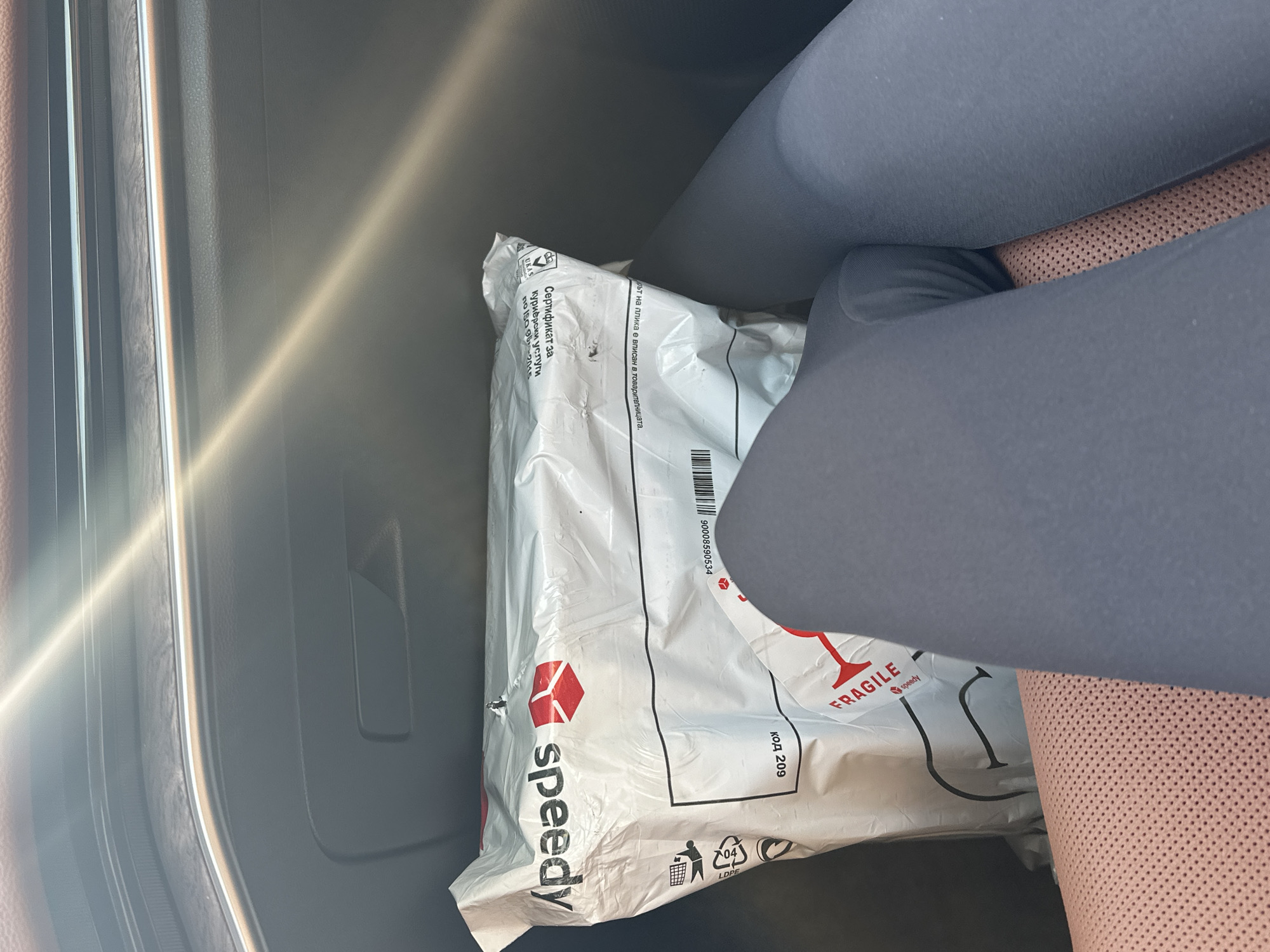 A Speedy package is visible on the floor of a car in Sofia, Bulgaria. The package is white and has red Speedy logo. There is also a "FRAGILE" sticker on the package. The car's interior is visible, including the seat and dashboard. The sun is shining through the windshield, casting a glare on the dashboard. The perspective is from the passenger seat, looking down at the package and the passenger's legs.