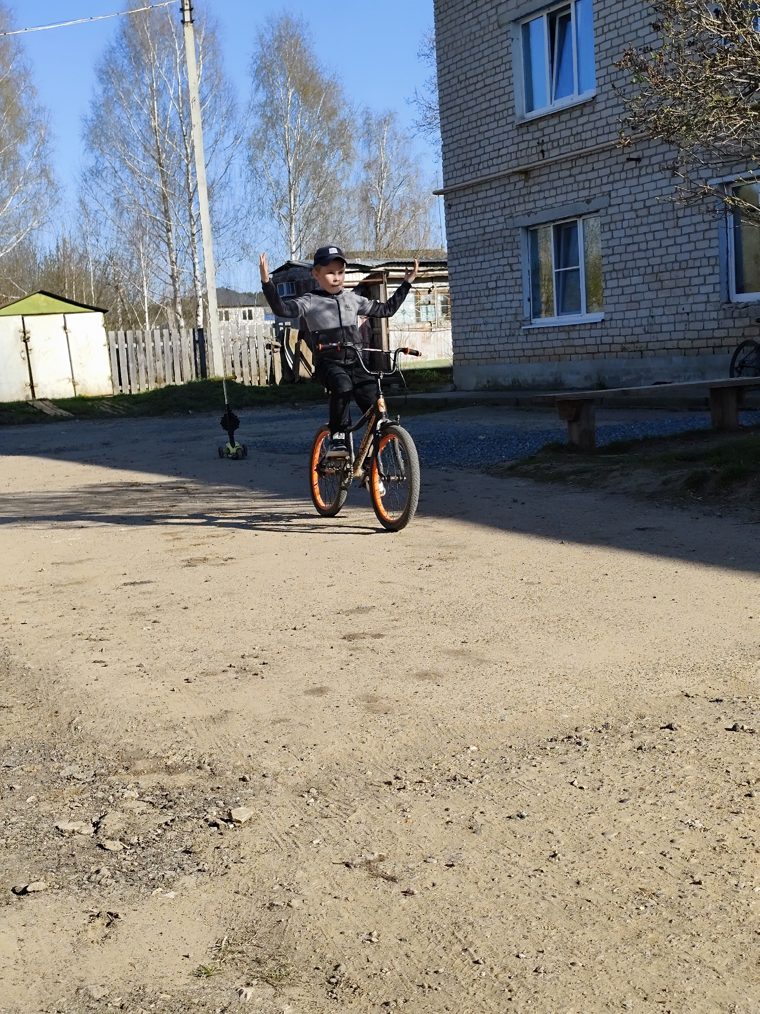 A young boy rides a bicycle on a dirt road in the city of Фоки, Russia. He is wearing a black cap, a gray and black jacket, and dark pants. His arms are raised in the air. A brick building is on the right side of the frame, and a shed and wooden fence are on the left. The road is dirt with patches of grass and exposed soil. In the background, bare trees are visible against a clear blue sky. A green children's scooter is to the left of the boy.
