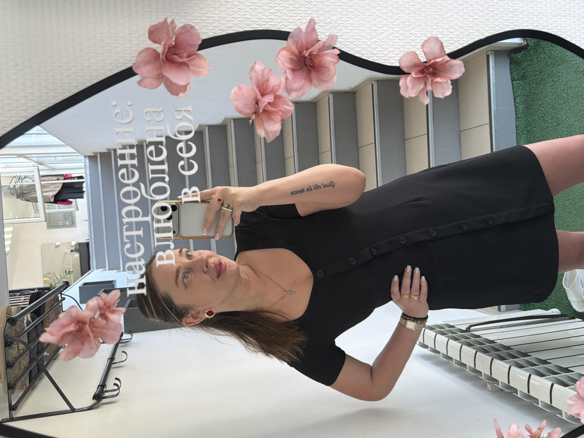 A woman is visible in a mirror taking a selfie. She has brown hair, blue eyes, and is wearing a black dress with buttons down the front. A gold necklace with a cross pendant is around her neck. She has a tattoo on her left arm. The mirror has pink flowers attached and an inscription in a Cyrillic alphabet. In the background, there are stairs, clothing racks, and a shoe rack. The location is the city of Chaikovsky, Russia.