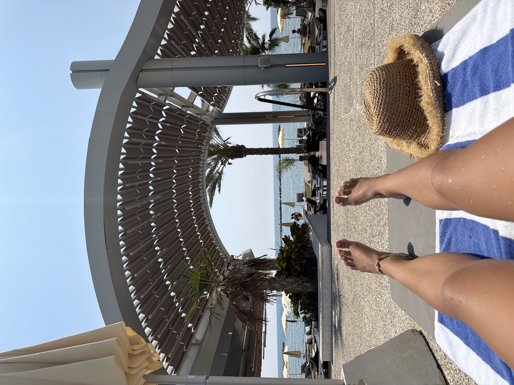 The image captures a first-person perspective of someone relaxing on a lounge chair by the sea in Oroklini, Cyprus. In the foreground, the individual's tanned legs and feet are visible, resting on a towel with blue and white stripes. A straw hat with a fringed brim lies on the towel next to the legs.
The background reveals a modern outdoor seating area with a distinctive curved, slatted canopy supported by grey pillars, providing shade. Beyond this structure, palm trees and lush green foliage are planted. Further in the distance, the calm, blue sea stretches out towards the horizon, dotted with several white umbrellas and lounge chairs set up on a beach or poolside area. A few people can be seen in the water and on the shore, enjoying the sunny day. The sky is clear and bright blue. The overall environment suggests a resort or hotel setting by the Mediterranean coast.