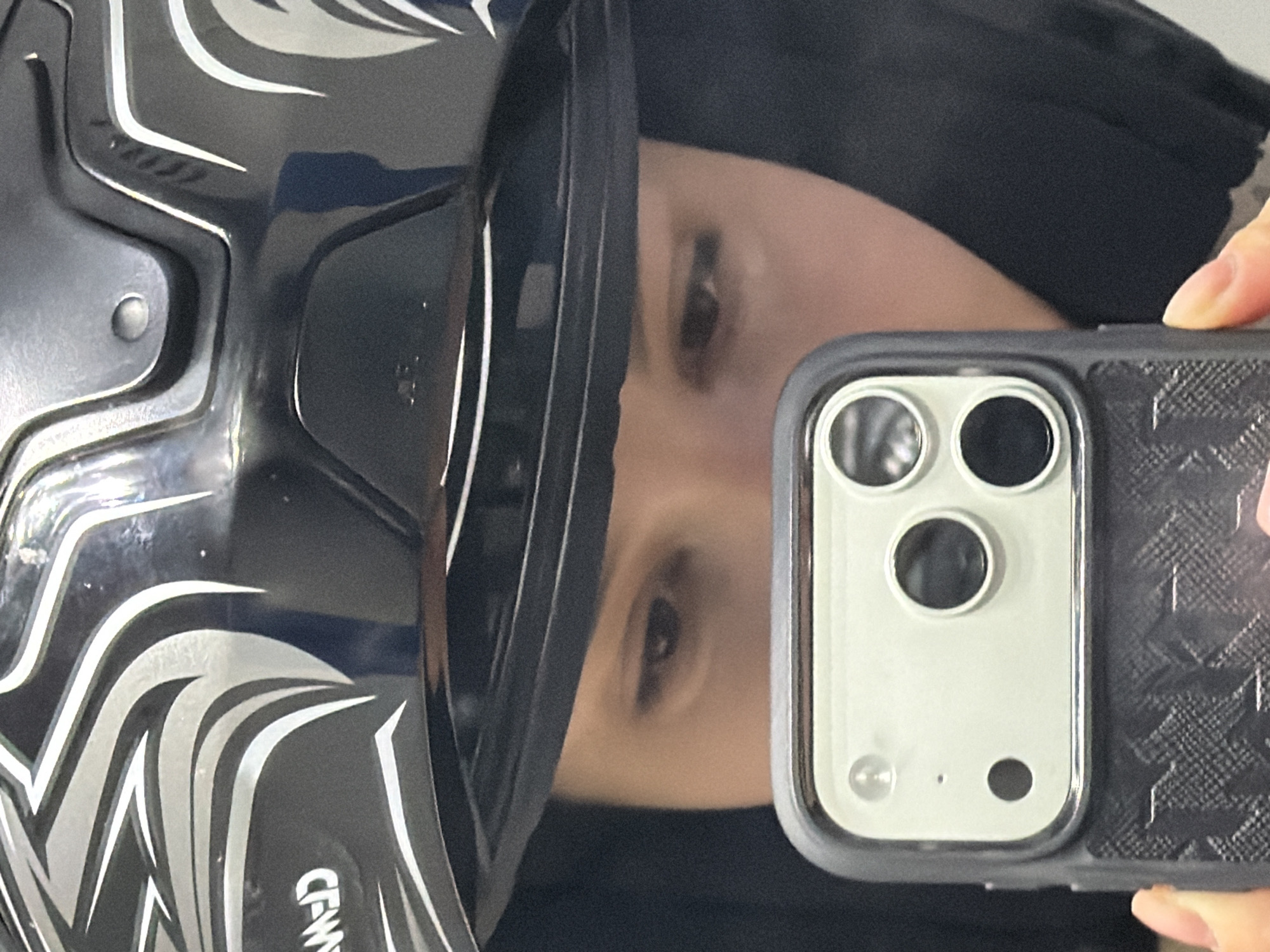 A close-up, selfie-style photograph shows a person's face partially visible in the reflection of a black and silver-patterned motorcycle helmet. The person is holding a mobile phone with a triple-lens camera and patterned case. The background is out of focus and dark. There's no information available to suggest a location in Moscow or even in Russia.