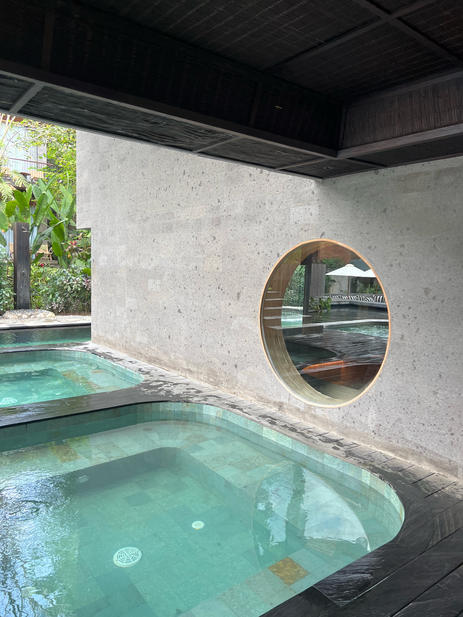 The image shows an outdoor spa area in Ubud, Indonesia, featuring multiple turquoise pools with tiled bottoms. The pools are surrounded by dark wooden decking, some of which is wet. A textured concrete wall with a circular window is prominently visible. Through the window, a larger pool area with lounge chairs and an umbrella can be seen. Lush green vegetation, including large-leafed plants, is visible in the background, suggesting a tropical setting. The ceiling above the spa area is made of dark, textured material, possibly bamboo or wood. No people are present in the immediate area of the pools.