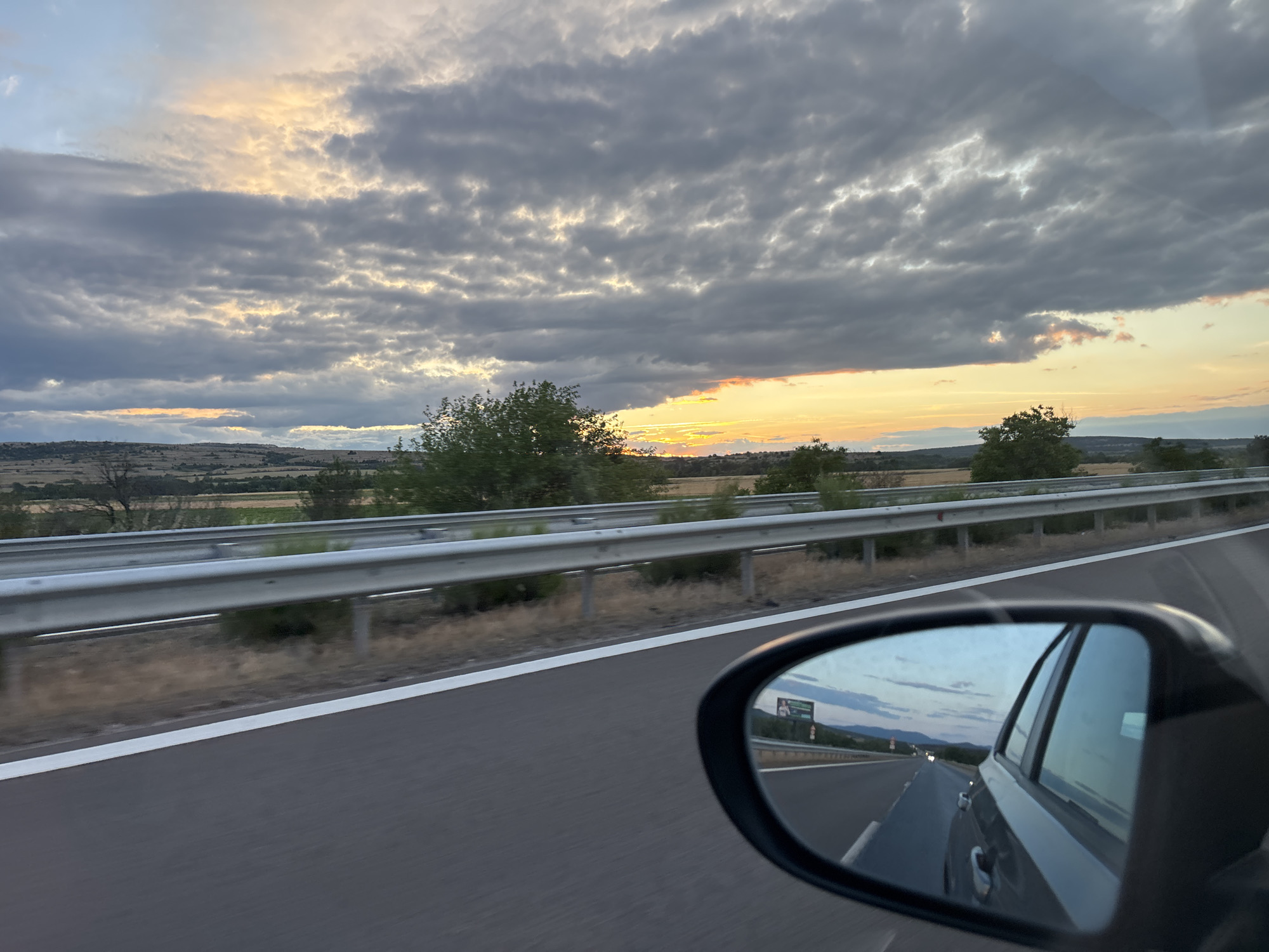 The image captures a view from a vehicle traveling on a highway, likely near Kalugerovo, Bulgaria, as suggested by the context. The scene is dominated by a dramatic sky filled with heavy, dark clouds interspersed with patches of bright, golden sunlight, indicating either sunrise or sunset. The highway runs diagonally from the bottom right towards the upper left, with a metal guardrail separating it from a dry, grassy verge and then fields. In the foreground, the side-view mirror of the vehicle reflects the highway stretching behind, with a few distant vehicles visible and a billboard to the left. The overall impression is one of movement and a vast, atmospheric landscape.