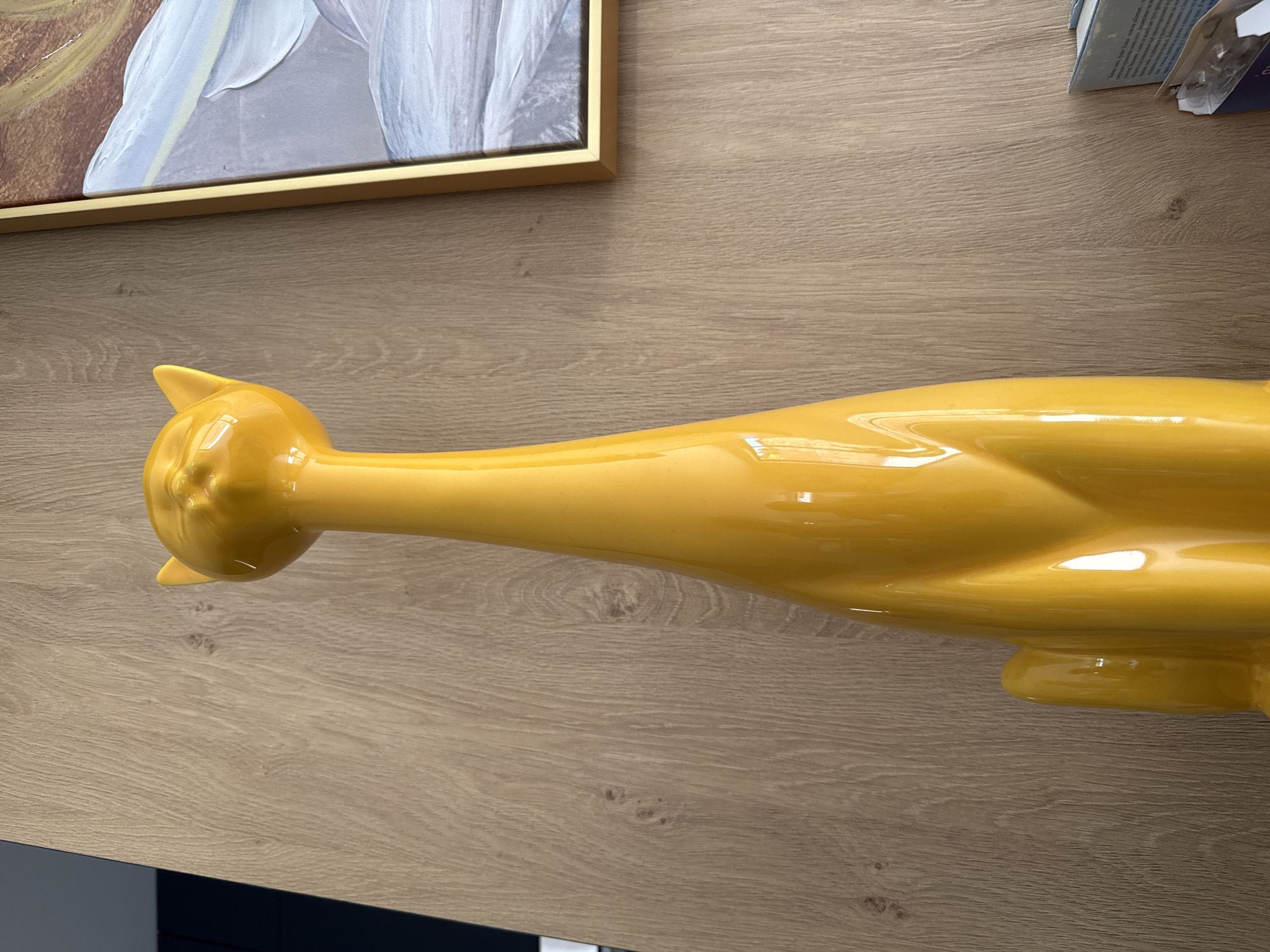 A medium-angle, eye-level shot depicts a yellow ceramic cat figurine laying on a wooden surface, positioned next to a framed painting. The cat figurine is oriented horizontally. The top of a stack of books is in the upper right. The lower-left edge is black. The image appears to be indoors. I cannot determine the specific location in Sofia, Bulgaria without additional information.
