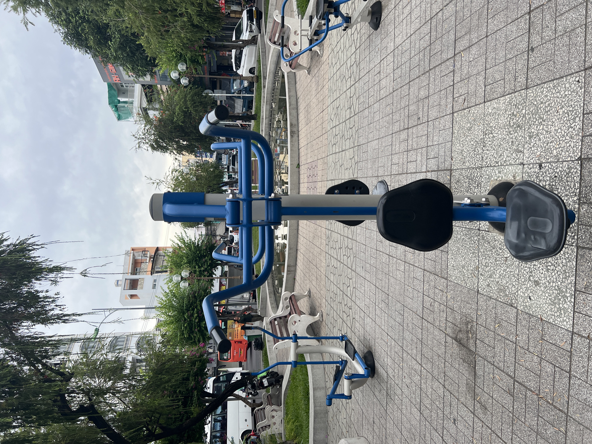 The image depicts an outdoor fitness area in a city park, likely located in Nha Trang, Vietnam, given the architectural style of the buildings visible in the background and the presence of a white bus with Vietnamese lettering. In the foreground, a blue and silver exercise machine with a black seat is centrally positioned. To its left, another similar machine with a white frame and blue accents is visible, along with a park bench. To the right, a portion of another exercise machine and a bench are seen. Behind the fitness equipment, a circular water feature with multiple small fountains is present, surrounded by a tiled pathway and low concrete planters with grass. Beyond the park area, a street scene unfolds with trees, various buildings, and vehicles, including a white bus, an orange bus, a motorcycle, a blue truck, and a white SUV. The sky is overcast.