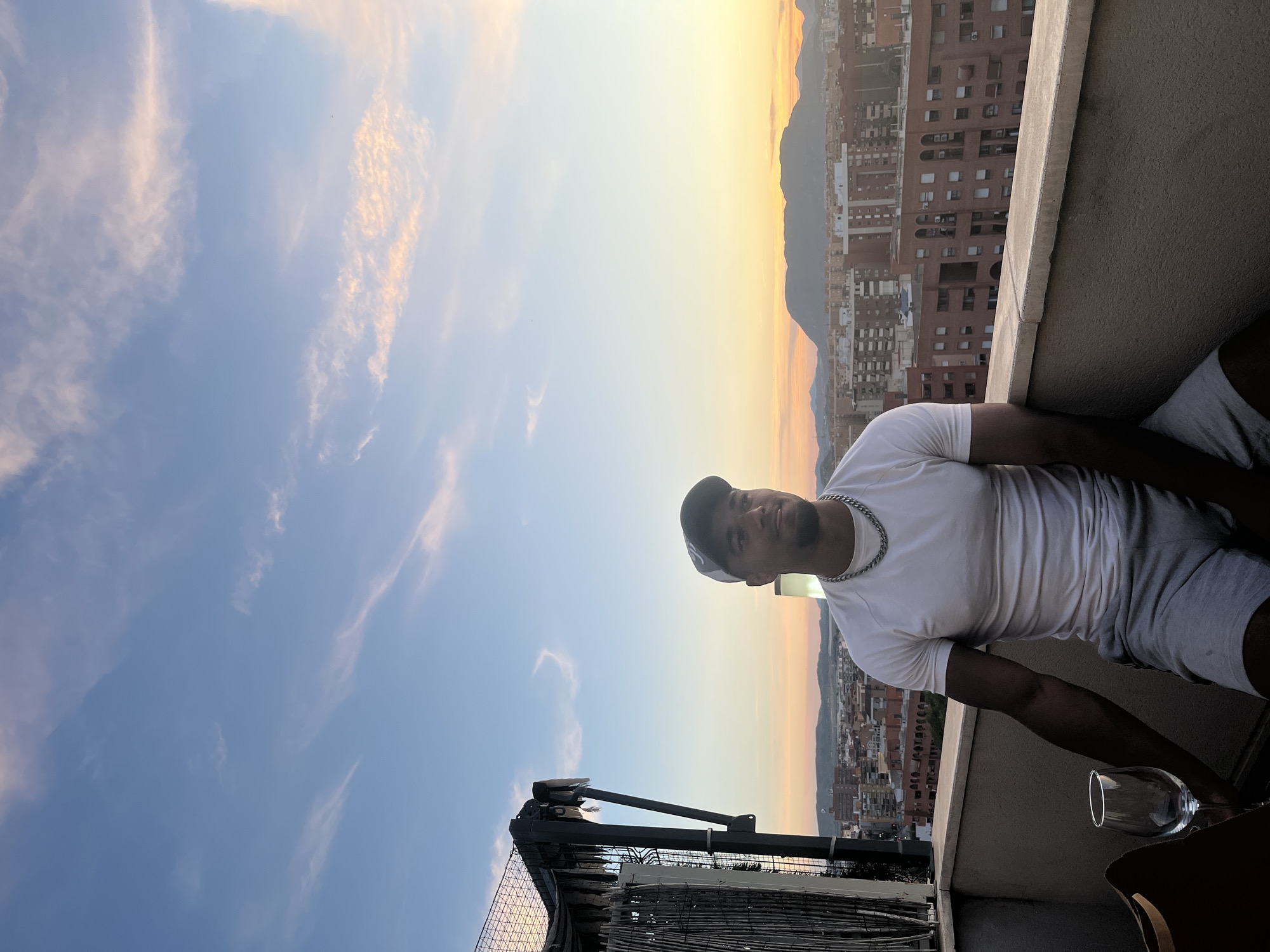The image shows a man on a balcony at sunset. The man wears a white t-shirt, shorts, and a cap and has a chain around his neck. He is looking at the camera. A wine glass is on the table near him. The balcony overlooks a cityscape with buildings and mountains in the background. The sunset creates a gradient of orange and yellow hues. Based on the buildings and mountains, the location appears to be Alicante, Spain.