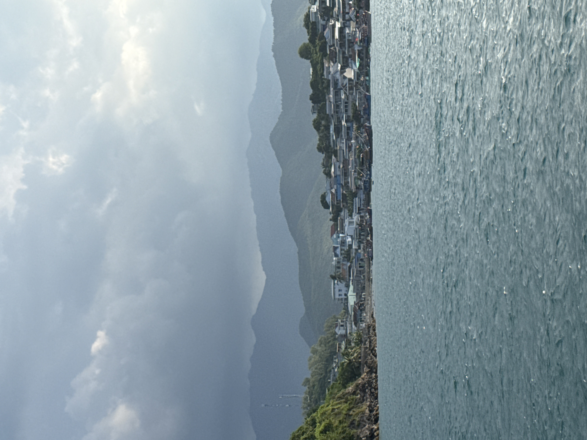 This image depicts a coastal village with a backdrop of lush, green mountains and a cloudy sky. The foreground is dominated by the turquoise waters of the sea, with gentle ripples reflecting the daylight. Along the shoreline, a dense cluster of buildings, typical of a Vietnamese coastal settlement, is visible. These structures are a mix of houses and businesses, with many featuring colorful roofs and visible antennas. Several fishing boats are moored or anchored in the bay, indicating a reliance on the sea for livelihood. The terrain rises behind the village, showcasing a steep, rocky incline covered in dense vegetation, leading up to layered mountain ranges that fade into the distance. The overall scene captures a vibrant, working community set against a dramatic natural landscape.