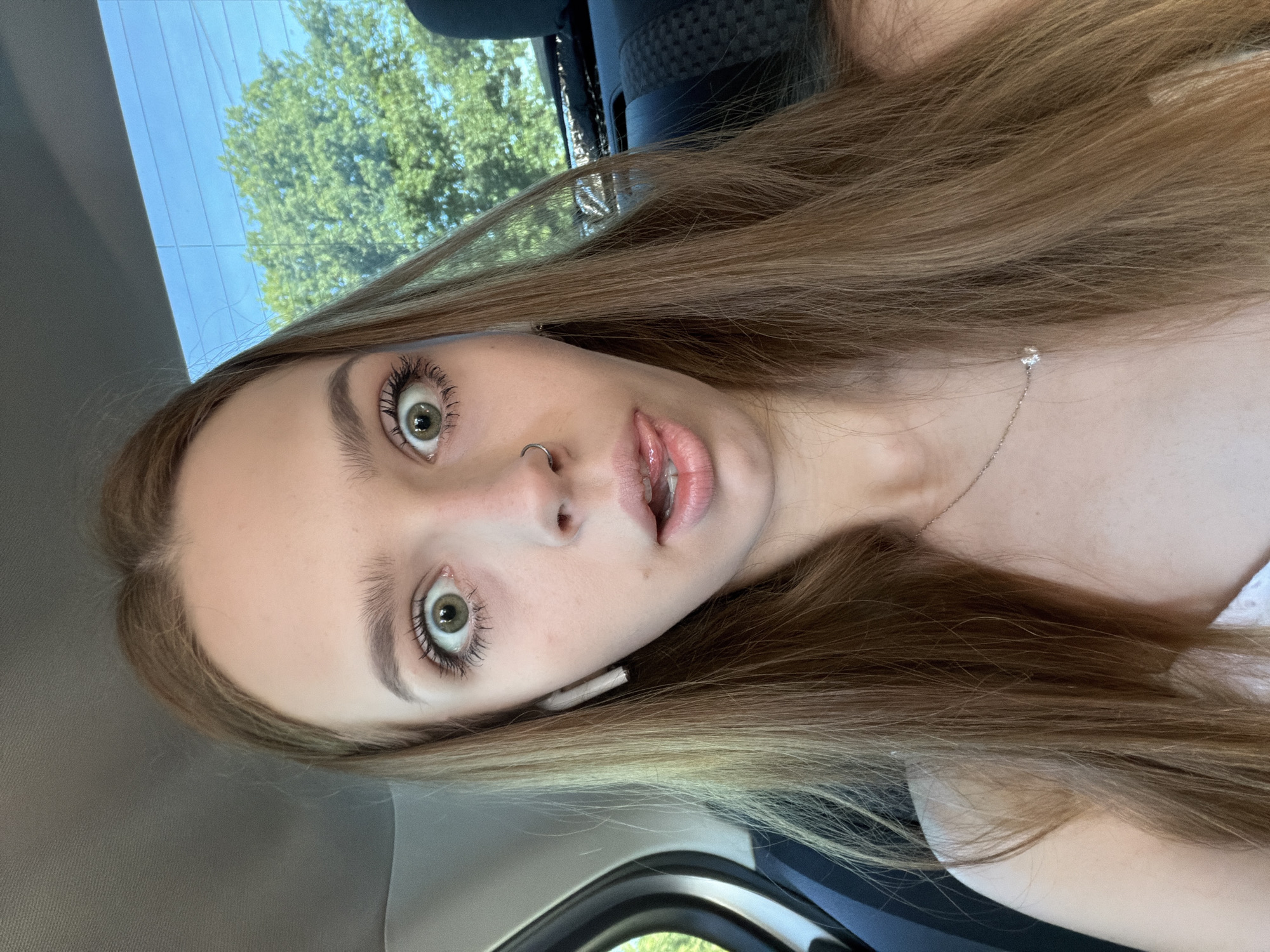 The media file is a close-up selfie of a young woman. She has long, straight, light brown hair that falls around her face. Her eyes are wide open, with green irises and dark, defined eyelashes. She is wearing subtle eye makeup and has a small nose ring. Her lips are slightly parted, revealing her teeth, and appear to be coated in a light-colored lip gloss. She is also wearing a delicate silver necklace with a small, clear pendant.
The setting appears to be inside a vehicle, as suggested by the gray fabric on the left side of the image and the rounded edge of what looks like a car window in the lower portion. Through the window, a bright blue sky and green trees are visible, indicating it is daytime and likely fair weather. The overall mood conveyed by her wide-eyed expression and parted lips could be surprise, curiosity, or excitement. There is no visible text in the media file.