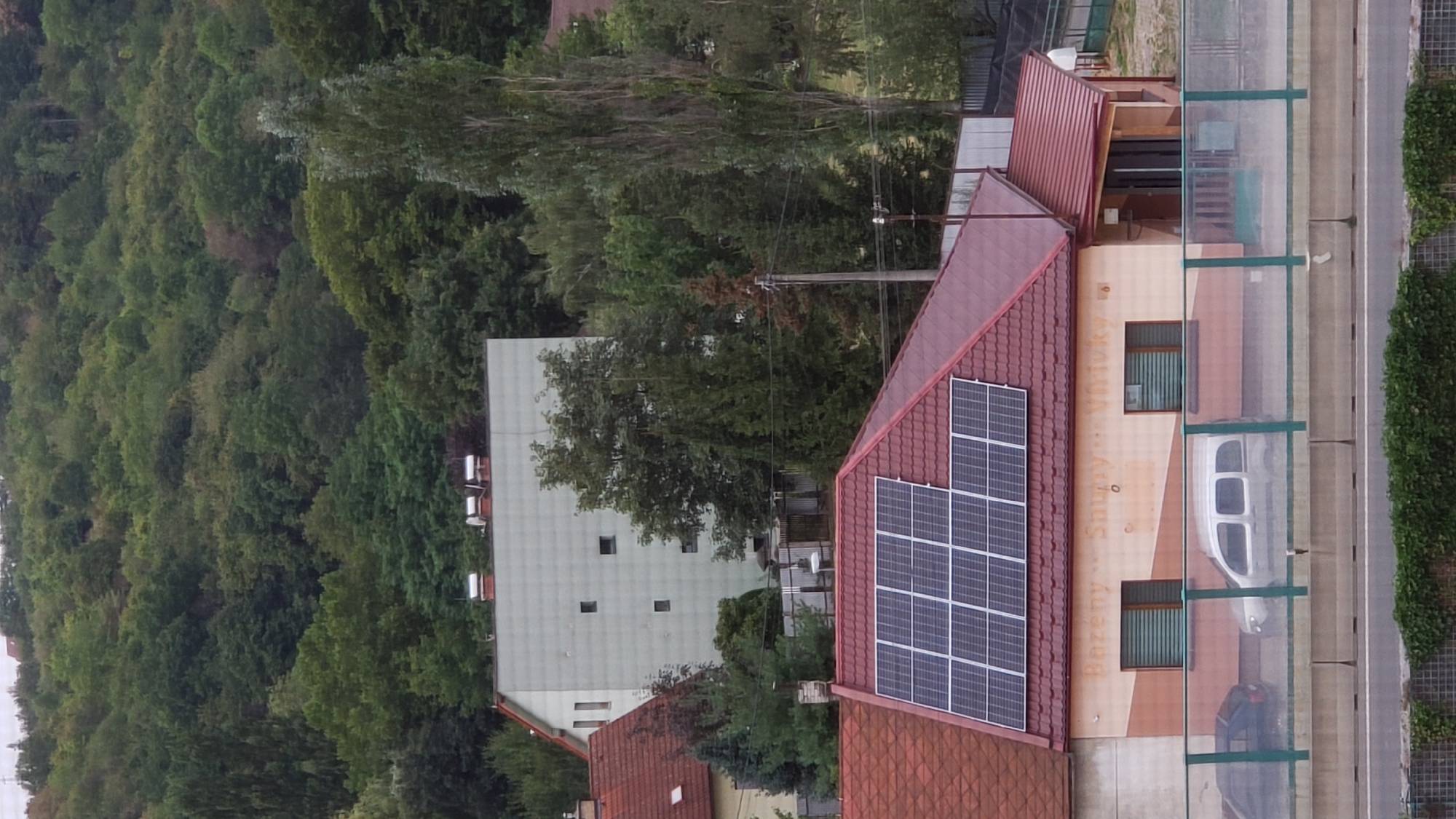 The image shows a residential area in Prešov, Slovakia. The foreground features a multi-story building with a red tiled roof covered in solar panels. A beige and brown facade has windows with closed blinds and text that reads "Bazény Snuby Vrsky". Parked in front of this building are two vehicles: a dark-colored sedan on the left and a silver minivan on the right. A transparent barrier with green accents separates the building's driveway from a road. Behind this building, a larger, pale gray building with a flat roof and small square windows is visible, surrounded by trees. The background is dominated by a densely wooded hillside. The overall scene depicts a residential neighborhood with modern infrastructure like solar panels integrated into the urban landscape.
