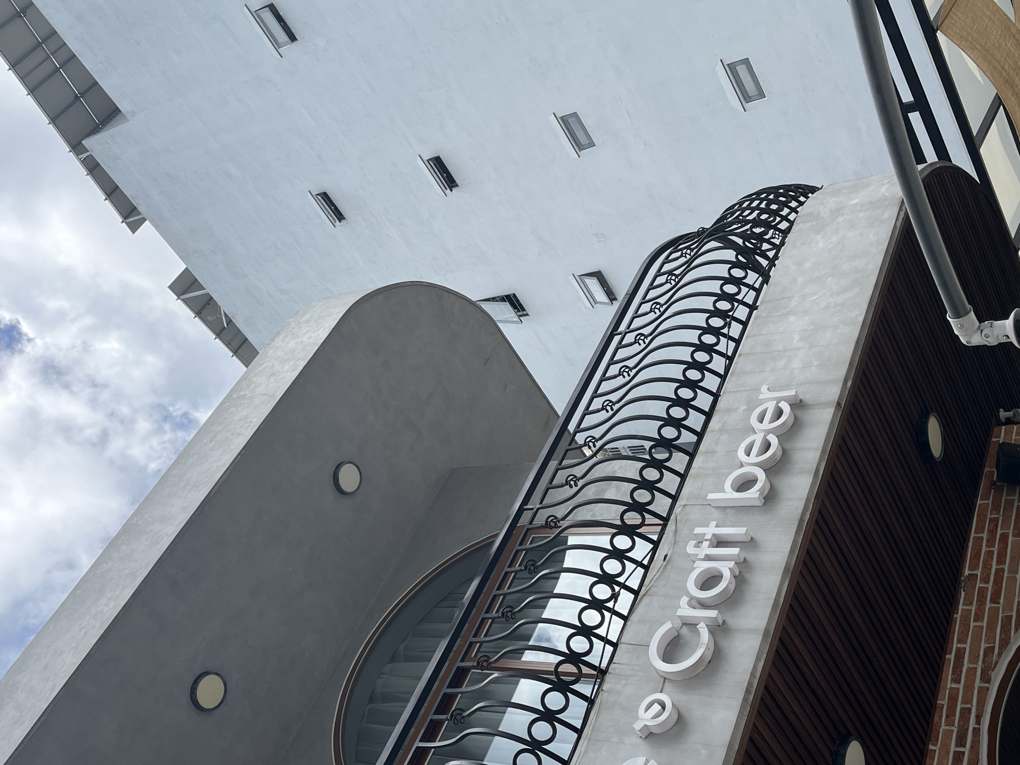 The image presents a dramatic, low-angle view of a modern building in Đà Nẵng, Vietnam, shot from below with a significant upward tilt. The bright, partially cloudy sky fills the upper left portion of the frame.
The architecture combines smooth concrete, wood, and brick. A prominent feature is a large, flat, light grey underside of an overhang or upper floor, seen from below, adorned with several small, rectangular windows or openings, some appearing slightly ajar.
On the left, a sleek, curved structure of smooth grey concrete rises, featuring embedded circular light fixtures. On the right, a multi-level facade showcases an elaborate black wrought-iron railing with wavy and circular patterns, suggesting a balcony or staircase. Below this railing, raised white lettering on a textured light grey surface clearly spells out "Craft beer," identifying the establishment. This section transitions into a facade of dark brown vertical wooden slats and a partial section of reddish-brown brickwork.
The perspective emphasizes the building's height and modern design elements. The scene is captured during daylight hours, indicated by the bright ambient light and the visible clouds.