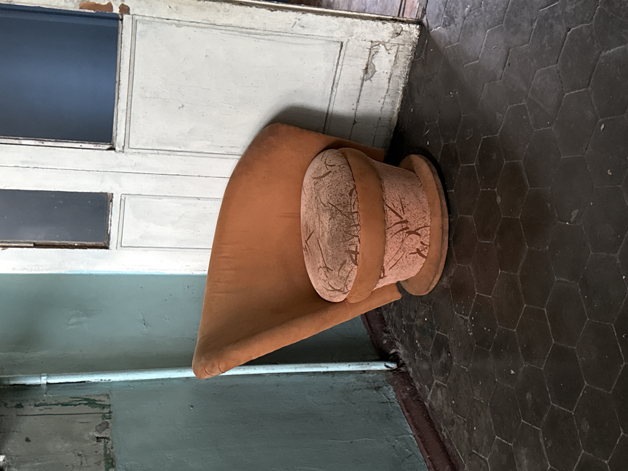 A vintage, burnt-orange upholstered chair with a rounded, petal-like back and a cylindrical base sits in a dimly lit corner. The seat is a lighter shade of orange with a textured pattern resembling dried leaves or branches. The chair rests on a circular base, which is placed on a dark, hexagonal-tiled floor. To the left of the chair, a teal-colored wall is visible, showing signs of wear and peeling paint, with a thin, light blue pipe running vertically along it. Behind the chair is a large, white wooden door with two panels, one above the other, each containing a dark pane of glass. The door also shows signs of age and wear, with chipped paint and visible wood grain. The environment appears to be an interior space, possibly an entryway or hallway, with a somber and aged aesthetic. There are no people visible in the image. The location is identified as Novorossiysk, Russia, suggesting this scene is within that city.