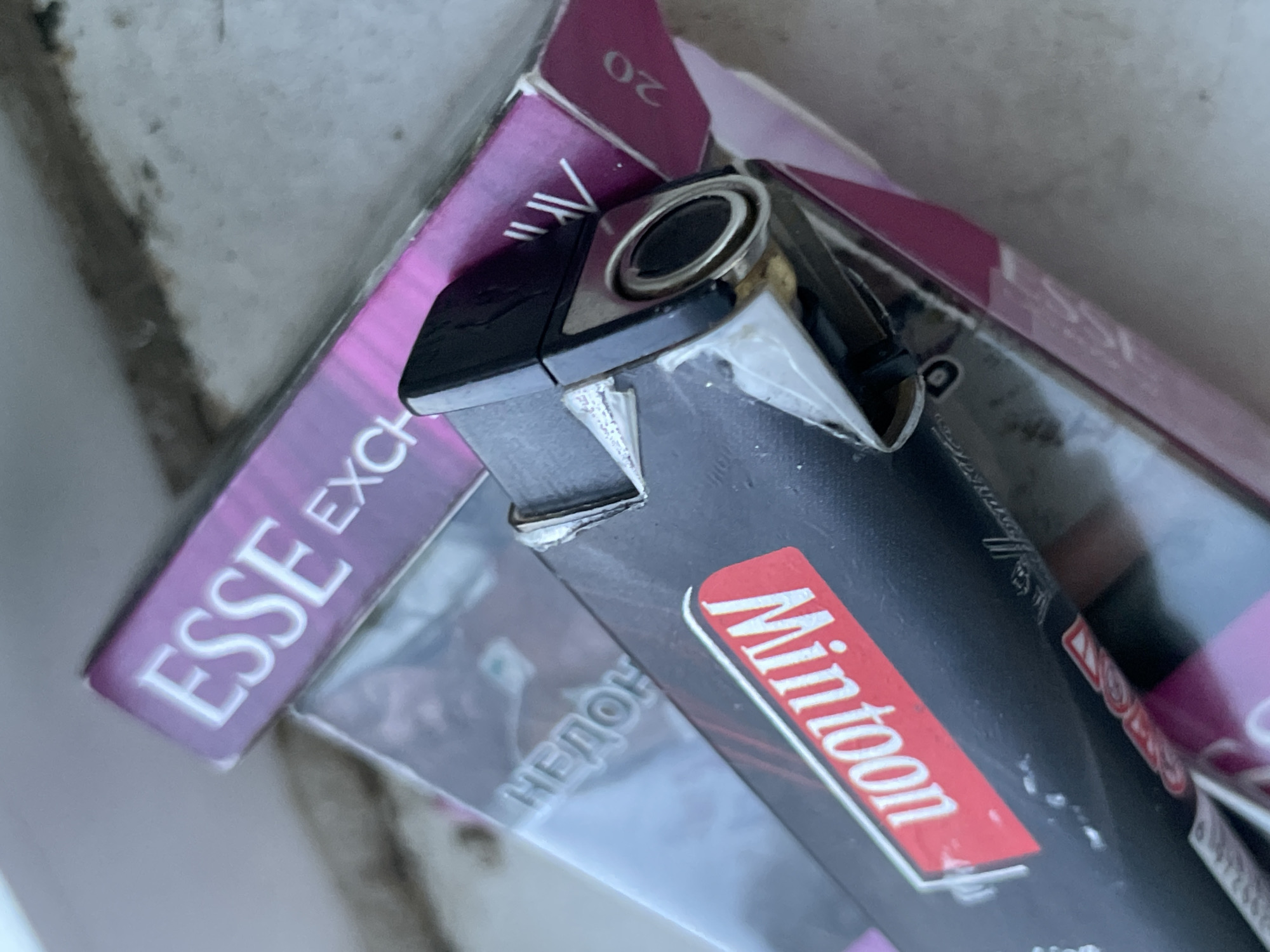 The image shows a close-up, top-down view of several partially obscured cigarette packs and a black lighter. The cigarette packs appear to be mostly pink and purple, with visible branding including "ESSE EXCH" on one pack and "Mintoon" on a red sticker on another. The lighter is black with silver trim and a circular silver-colored opening. The background is a textured, light-colored surface that appears to be concrete or stone. The overall scene suggests an outdoor setting. There is no identifiable information or context within the image that places it in the city of Aktau, Kazakhstan, or any other specific location. The image is purely a visual record of the objects presented.