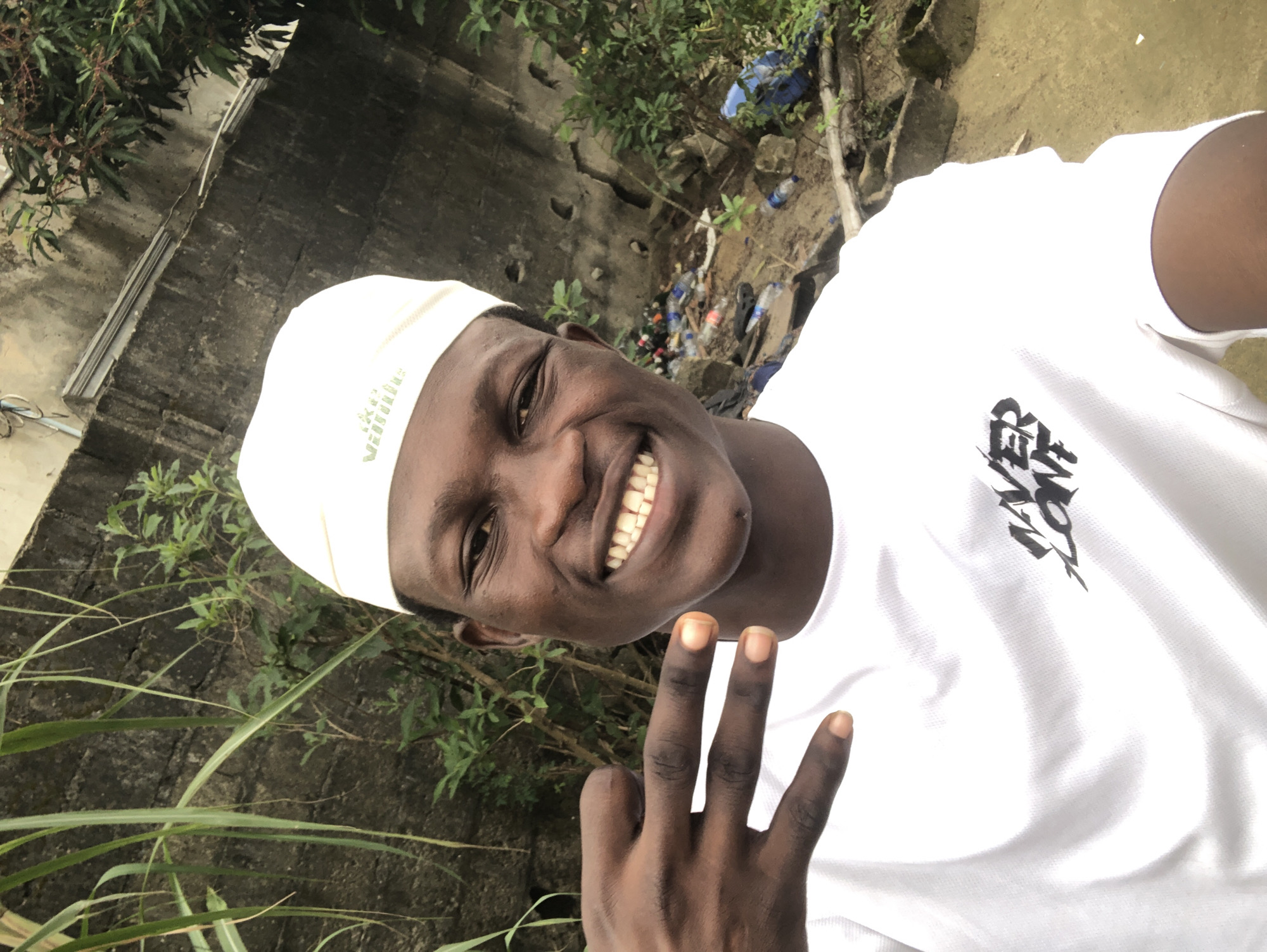 In Ado, Nigeria, a young Black male is captured in a selfie taken outdoors. He is wearing a white t-shirt with a black graphic design and a white skullcap. The man has a wide smile, revealing his teeth. His left hand is raised, with his index and middle fingers extended in a peace sign.
The background reveals a somewhat neglected outdoor environment. A rough, textured wall is visible, possibly made of concrete or stone, with patches of moss or discoloration. Lush green vegetation, including tall stalks of grass and leafy branches, grows in front of and around the wall. To the right of the man, the ground appears to be dirt or mud, and there is visible debris, including plastic bottles and other refuse, suggesting an area that may not be well-maintained. The overall impression is of an urban fringe or a less developed part of the city.