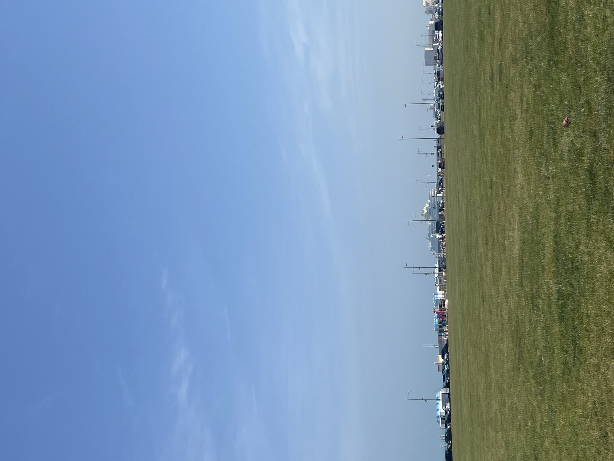 An outdoor, daytime, eye-level, wide shot shows a vast, green, manicured lawn in the foreground. In the middle ground, a parking lot is visible, filled with numerous cars and recreational vehicles. Beyond the parking lot, a cityscape unfolds, characterized by various buildings, including what appears to be a ferry terminal or port with large ships docked. Lampposts are scattered throughout the scene. The background is a clear, pale blue sky. The location is the grassy area near the port of Dieppe, France.