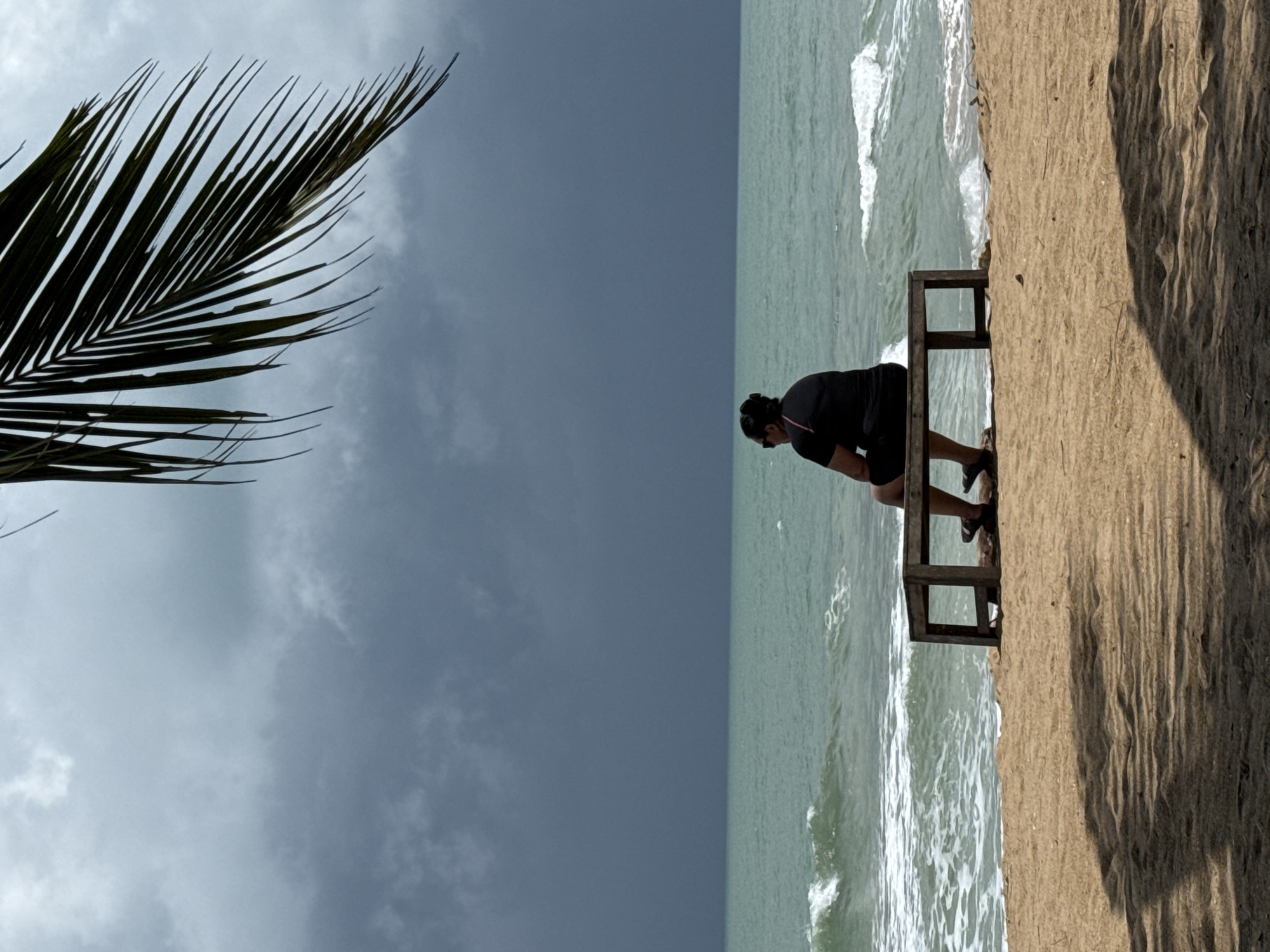 A person sits on a wooden bench on a sandy beach in Ban Khuk Khak, Thailand, facing a turbulent sea under a dark, cloudy sky. The person, seen from behind, wears a black t-shirt and shorts, with dark hair pulled back and sunglasses. They are seated on a simple, low wooden bench. The sea is a greenish-blue with white-capped waves. The sky is overcast with heavy, dark clouds, suggesting an impending storm. A palm frond hangs from the upper right corner of the frame, casting a shadow on the sand in the foreground, which also displays a distinct shadow pattern.