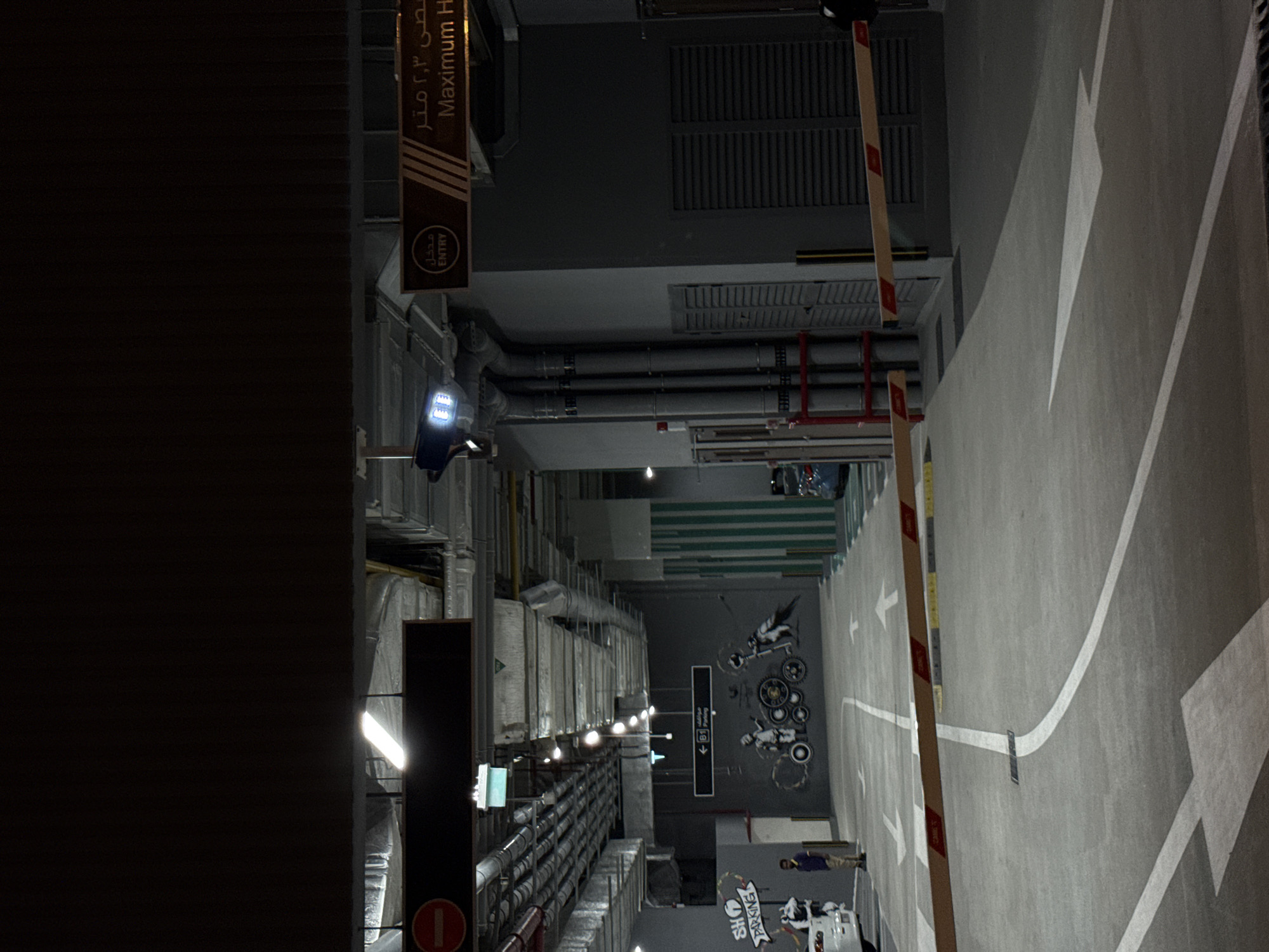 The image depicts the interior of a dimly lit parking garage, likely in Dubai, United Arab Emirates, indicated by signage. Overhead, a complex network of pipes and ventilation systems is visible, illuminated by fluorescent lights. On the right side of the image, a sign displays "Maximum H" with Arabic text above, suggesting a height restriction. A security barrier arm, marked with red and white stripes, is positioned across the lane. To the left, a black sign with a red circle and a horizontal bar, signifying a prohibition, is present. Further left, another sign points towards "B1 Parking." A mural featuring stylized figures and bicycles adorns the wall. A man in casual clothing stands near a white car on the far left. The floor is marked with directional arrows and white lines indicating parking bays. The environment is industrial and utilitarian, typical of a multi-level car park.