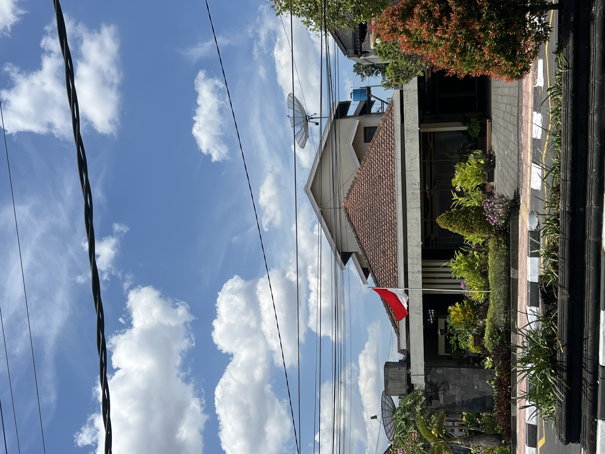 The image captures a view of a building in Yogyakarta, Indonesia, on a bright, sunny day. The sky is a vibrant blue, dotted with fluffy white cumulus clouds. Several power lines traverse the upper portion of the frame. In front of the building, an Indonesian flag, red and white, flies from a flagpole. The building itself has a tiled roof and appears to be of commercial or institutional design. There are satellite dishes visible on one of the rooftops, and a blue water tank is also present. The foreground shows a well-maintained garden with various types of plants and flowers, bordered by a paved walkway and a street. The overall scene suggests a daytime setting, likely in an urban or suburban area of Yogyakarta. No people are visible in the image.