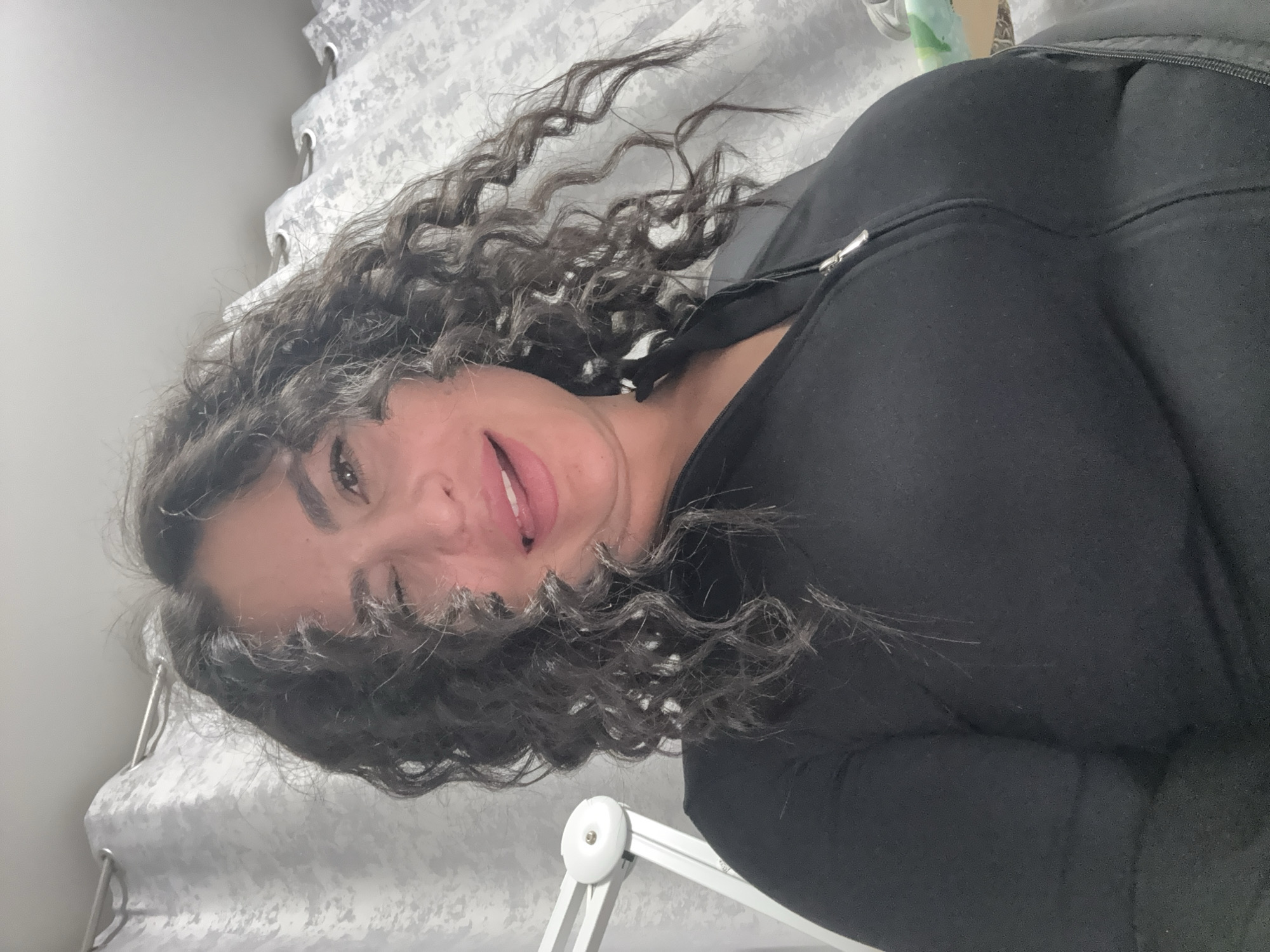 The image shows a woman with curly dark hair wearing a black zip-up jacket. She is sticking out her tongue and winking. The background includes a textured white curtain. A white desk lamp is partially visible in the bottom left corner. There is no evidence to suggest the image was taken in Kauchuk Stantsiya, Russia.