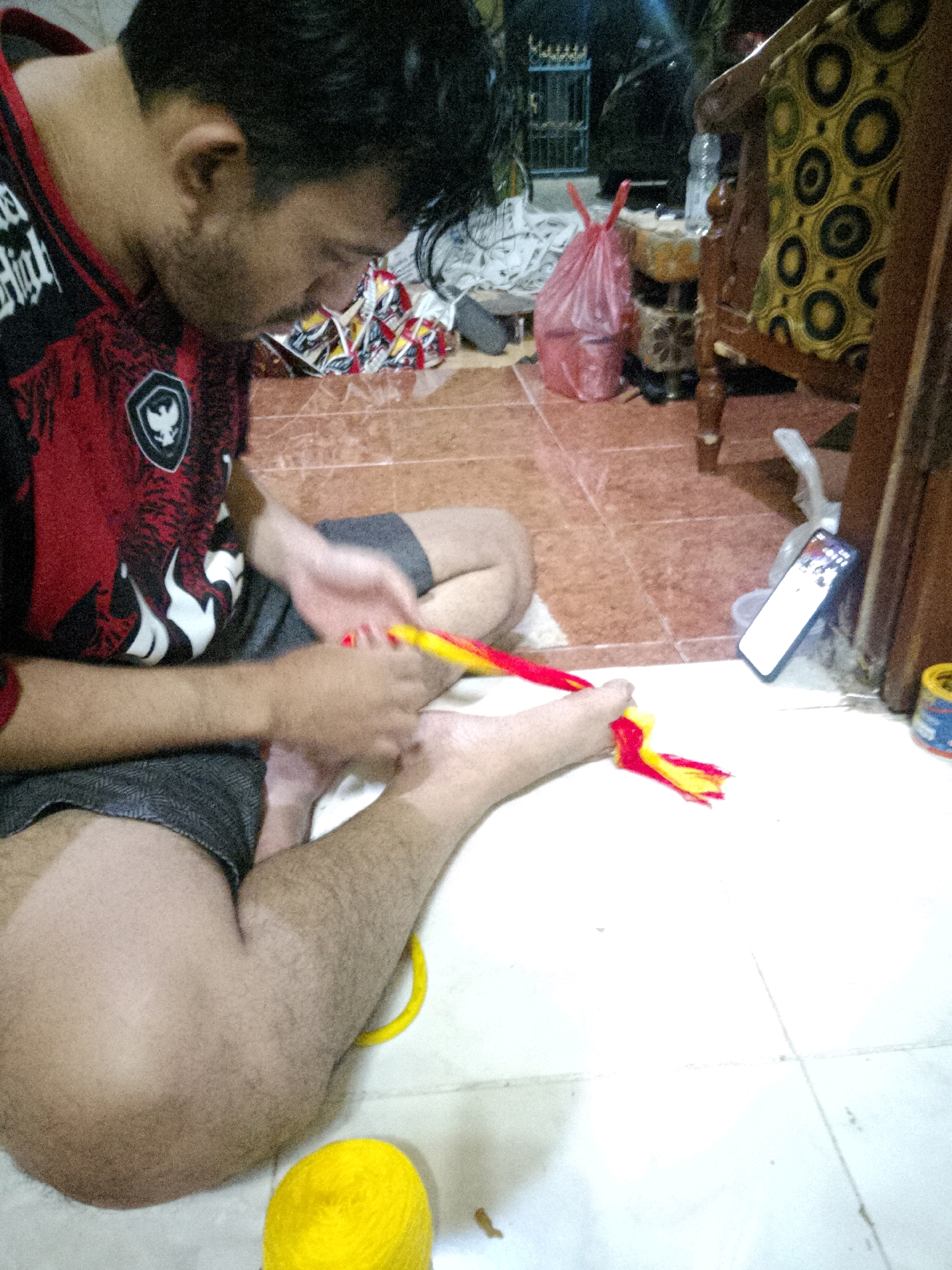 In Gampengrejo, Indonesia, a person with dark hair and a beard, wearing a red and black shirt and dark shorts, is sitting on a tiled floor. They are focused on their feet, where they are manipulating two colorful braided cords, one red and yellow, and the other red and yellow. One cord is wrapped around their left foot. A ball of yellow yarn is visible on the floor near their feet. In the background, a wooden chair with a patterned cushion and a pink plastic bag are present. A smartphone is propped up on a surface, displaying a bright screen. The environment appears to be indoors, with the floor made of reddish-brown tiles and a white tiled area.