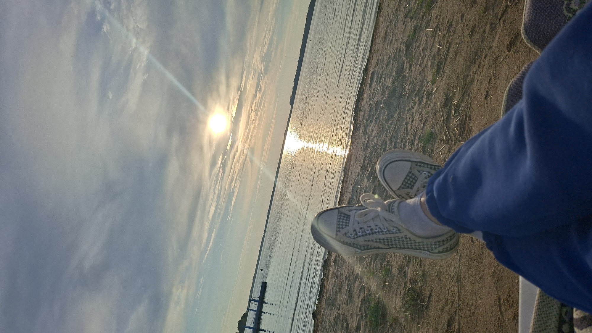 An outdoor, eye-level, medium shot captures a person's legs and feet relaxing on a sandy beach by a body of water. The person is wearing blue sweatpants and white sneakers with a checkered pattern. The sun is setting over the water, casting a warm glow and creating a shimmering reflection on the surface. The sky is partly cloudy with streaks of light. In the distance, there is a dock and some faint landmass. The scene is serene and evokes a sense of peace and relaxation. The location appears to be the beach area of Myadzyel Lake, Myadzyel, Belarus, given the characteristic landscape of the region.
