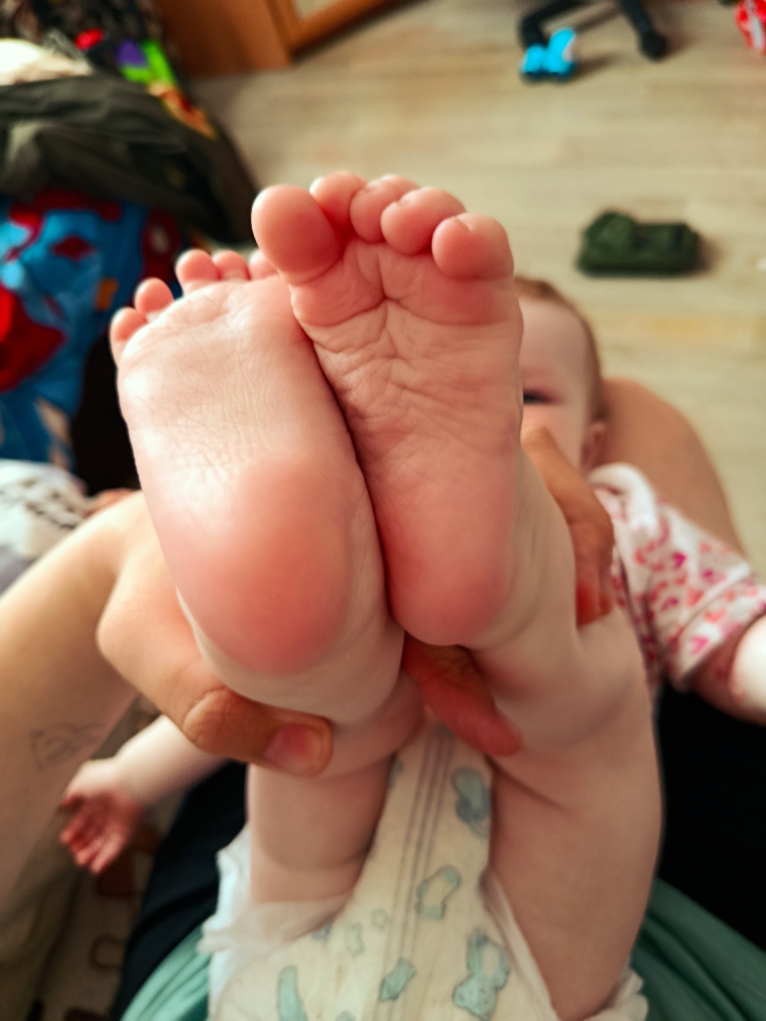 A close-up indoor shot focuses on a baby's feet held up by a hand. The soles of the feet are prominently displayed, with tiny toes visible. The baby is wearing a diaper patterned with blue figures. The baby's partial face, including an eye, is visible behind the feet. The person holding the baby is wearing a pink and white floral print top. The background includes a wooden floor with various objects out of focus, including what appears to be a blue toy. No elements in the image indicate the location is Фоки, Russia.