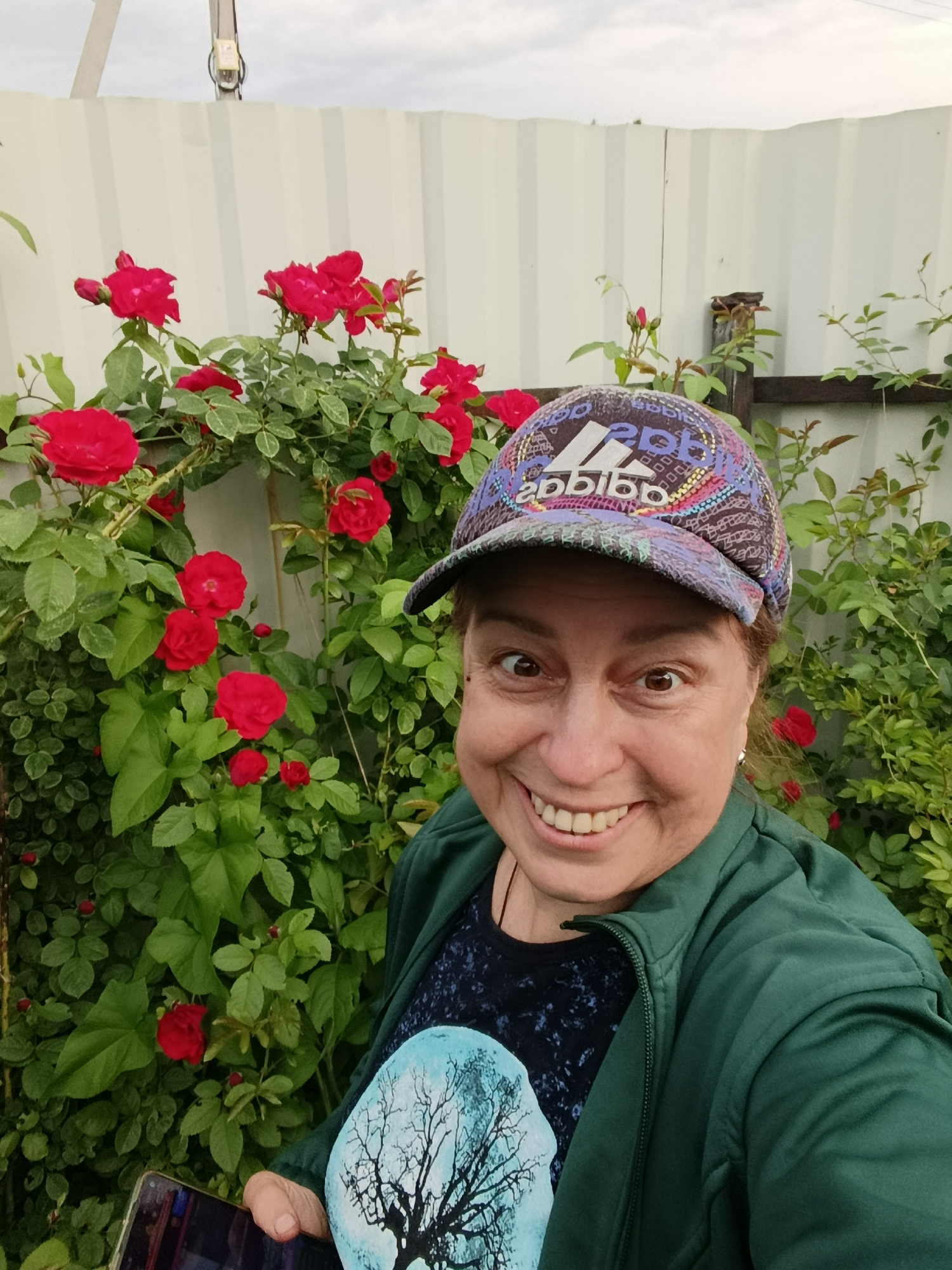 A woman is taking a selfie in a garden with red roses and a white fence. She is wearing a green jacket, a dark t-shirt with a tree design, and a colorful baseball cap. The woman is smiling broadly at the camera. The background shows a cloudy sky and greenery. It is unknown if this location is in the city of Ver, Russia.