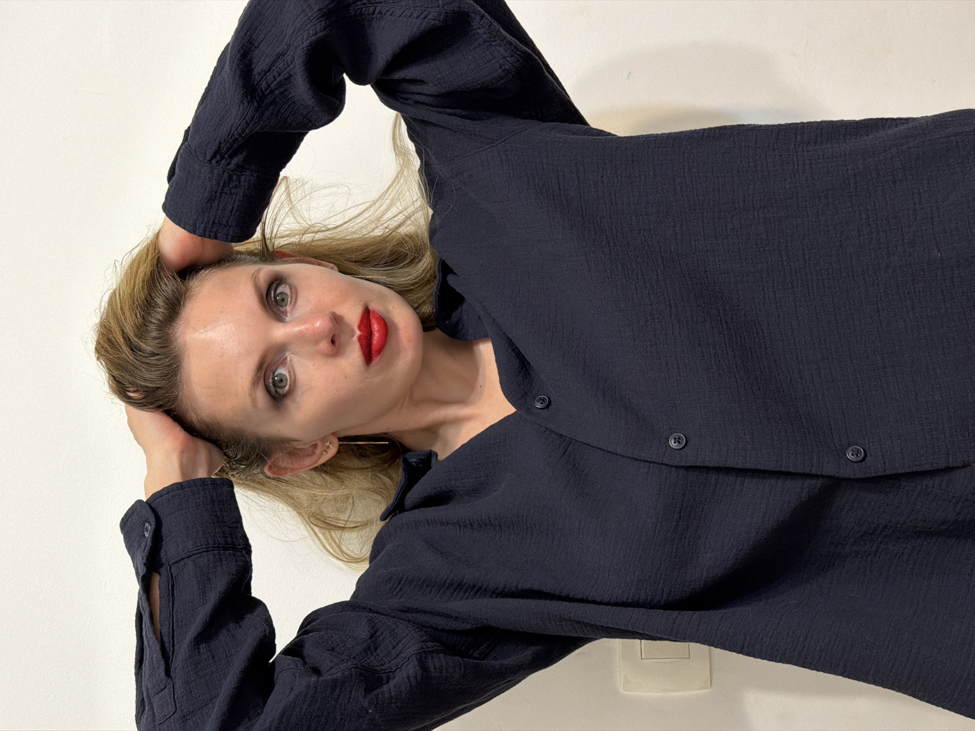 A woman with blonde hair and red lipstick is reclining, wearing a dark-colored, long-sleeved button-up shirt. She is holding her head with both hands. The background is a plain white wall. A light switch is visible in the lower right corner. There's no information to suggest this is in Budva, Montenegro.