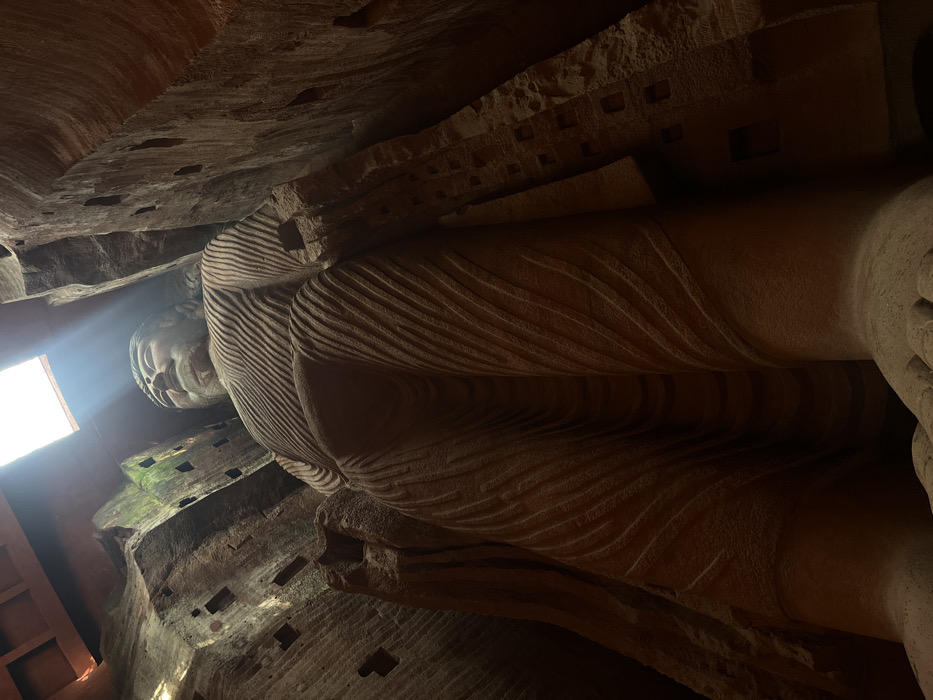 This image captures the colossal Leshan Giant Buddha, located in Shizhong District, Leshan, Sichuan Province, China, viewed from a low-angle perspective looking upwards. The immense stone-carved statue, depicted in a standing posture, features a serene facial expression with a prominent nose and tightly curled hair. Its robe is rendered with numerous horizontal, undulating carved lines suggesting drapery. The Buddha is flanked by massive, rough-hewn rock walls, carved directly into a cliff face or grotto, which display multiple rectangular cutouts or recesses. Bright natural light emanates from a rectangular opening positioned directly above the Buddha's head, casting distinct rays downwards and illuminating the upper portions of the statue and surrounding rock, while the lower body remains in shadow. No other individuals or specific objects are present within the immediate foreground.