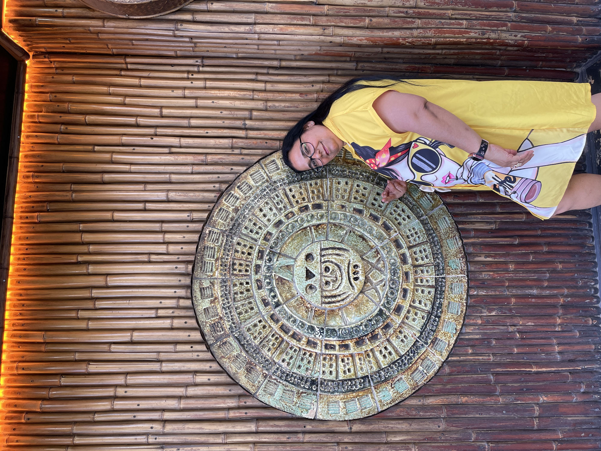 A woman with long black hair, glasses, and a yellow dress with a cartoon print is standing in front of a bamboo wall with a circular stone carving. The stone carving appears to be an ancient calendar or artifact. The bamboo wall has orange lighting along the top edge, creating a warm glow. The woman is smiling and looking towards the camera, with her left hand resting on the stone carving. The scene is likely an outdoor or semi-outdoor location, possibly a restaurant or cultural attraction.