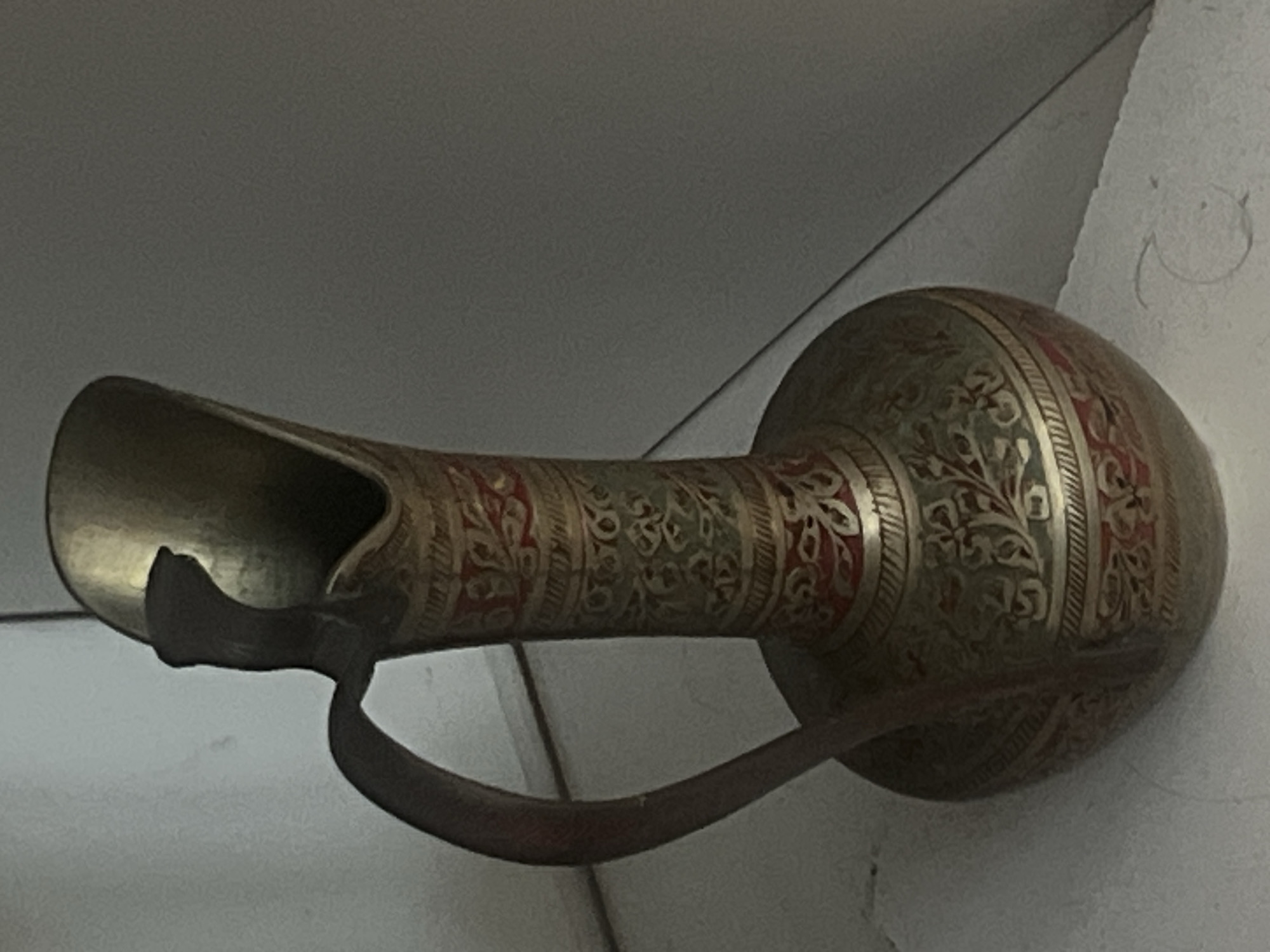 An intricately decorated brass pitcher or ewer stands upright, positioned slightly off-center to the left. The vessel features a tall, slender neck that flares into a rounded, bulbous base. Its surface is adorned with elaborate patterns, including floral and foliate motifs, etched or embossed into the metal. Bands of dull red and a muted greenish-blue enamel or paint contrast with the metallic brass, creating distinct horizontal segments around the body of the pitcher. The top portion of the vessel is plain brass, forming an elongated, open spout. A single, curved handle extends from just below the spout to the shoulder of the pitcher. The object is situated on a light-colored, flat surface, possibly a shelf or tabletop, which meets a vertical, light-colored wall at a right angle, forming a corner in the background. Subtle shadows are cast on the right side of the pitcher and the background wall. No people are present. This scene is located in the city of Casablanca, Morocco.