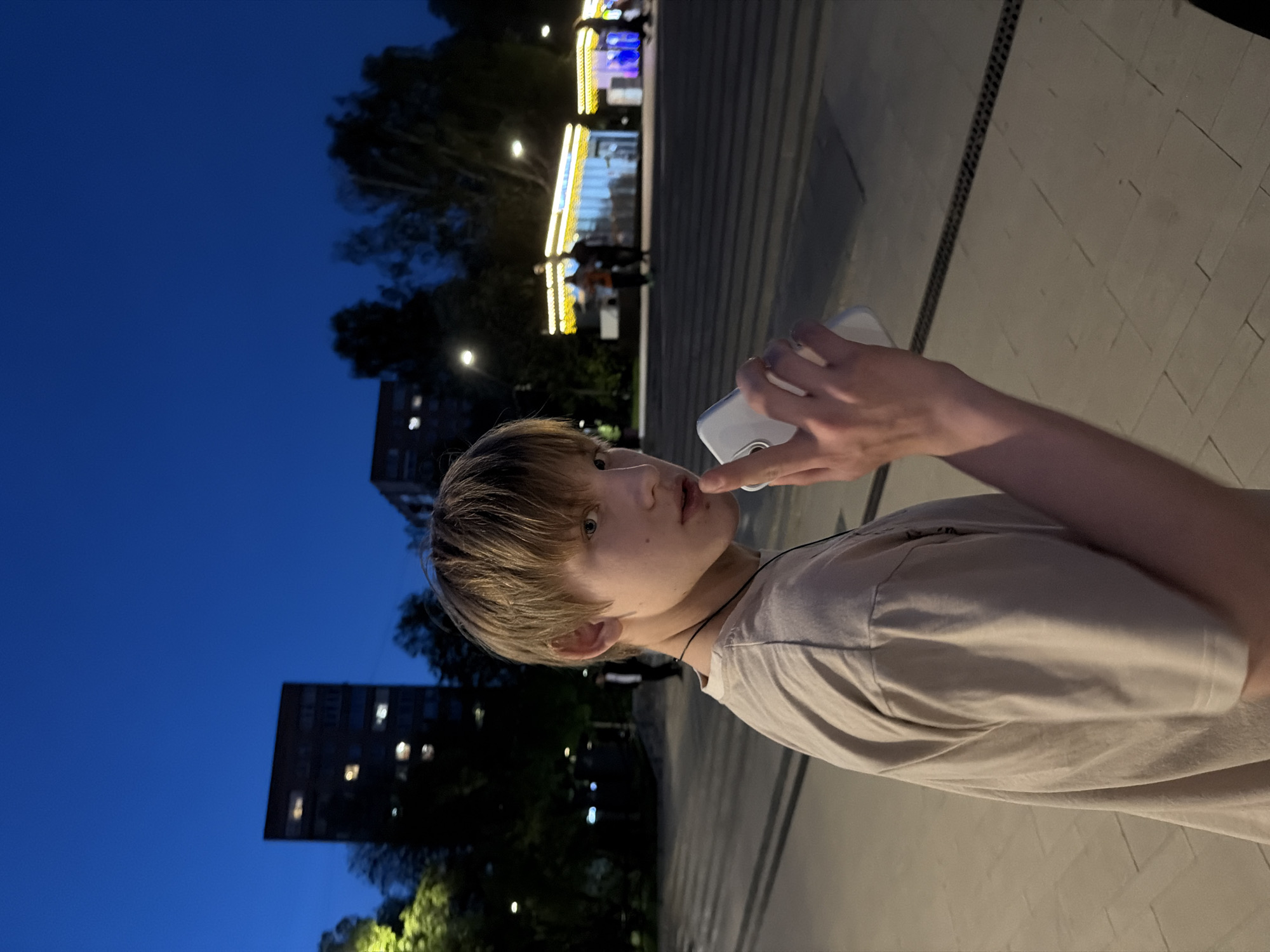 A young person with short, light brown hair and fair skin is holding a white device, possibly a phone, near their mouth. They are wearing a light-colored t-shirt and a thin black necklace. The person appears to be outdoors at dusk or night. In the background, there is a building with lit windows and trees. There are streetlights and a structure illuminated with yellow and blue lights, possibly a market or amusement ride. The foreground consists of light-colored pavement. Based on the background landmarks and the style of architecture, the location is potentially Izhevsk, Russia.