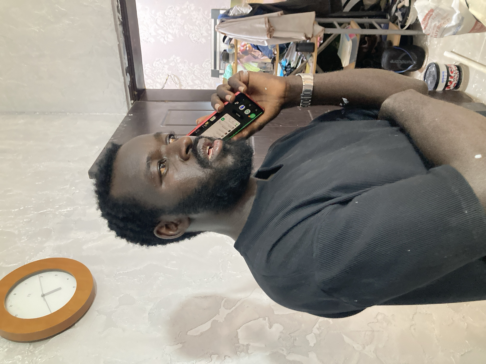 A man is captured in a close-up, upright portrait, seemingly engaged in a phone call indoors in Jos, Nigeria. He is a dark-skinned individual with a full beard, wearing a black ribbed shirt and a silver wristwatch on his left wrist. White specks, possibly paint, are visible on his arm and shirt. His mouth is slightly open as if speaking, and his eyes are looking upwards and slightly to the right, focused on something beyond the frame.
He holds a red-framed smartphone to his left ear, its black screen displaying several app icons, including a visible portion of "WhatsApp," and emits a green glow from its right edge. To his lower left, an oval-shaped wooden clock with a white face and simple black hands indicates approximately 2:11. The surrounding white walls and floor appear textured or splattered with a lighter substance, possibly paint, suggesting recent work or a disheveled environment.
In the cluttered background, a rack or shelf holds various items. A white bag with the partially visible text "ORIGINAL" and "BUY BEST" hangs alongside other objects. A black cylindrical container with a red and white label, featuring the numbers "999," is also present. The wall behind the man features a faint floral pattern. The overall scene suggests an ordinary moment, possibly during a break from an activity that involves paint or similar materials, in an indoor setting.