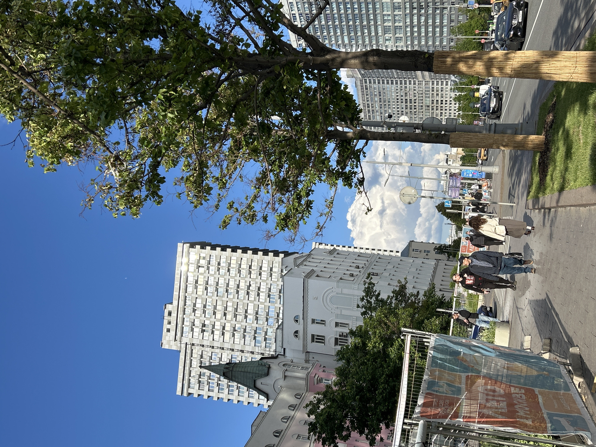 The image depicts a street scene in a city. Several buildings of various styles are visible, including a tall, modern building with many windows, a white building, and a pink-accented building with a green roof. People are walking on the sidewalk. Trees are present, some with trunks wrapped in protective material. A street with cars is seen to the right. A construction fence with a large poster is visible in the foreground. The sky is blue with some clouds.