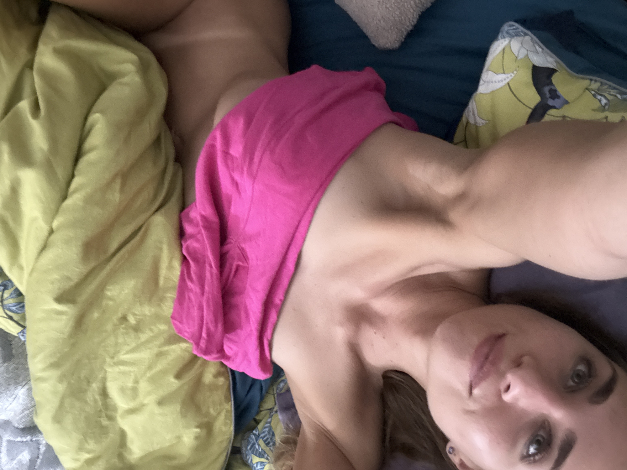 A woman is lying on a bed with her head to the left. She is looking directly at the camera with her eyes open and her mouth slightly open. She has blonde hair and light skin. She is wearing a pink top that is pulled up to cover her chest. The bed is covered with a yellow and green patterned comforter. There are also some pillows on the bed, one of which is gray and fuzzy. The room appears to be dimly lit. The image was taken in an unknown location in Russia.