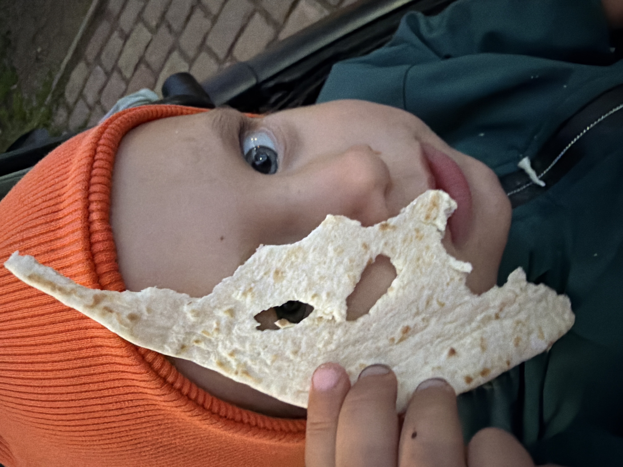 The scene, located in Chaikovsky, Russia, features a close-up of a young child, approximately toddler age, viewed from the chest up. The child is wearing an orange ribbed knit beanie and a dark teal zippered jacket. Their left hand holds an irregularly torn piece of light-colored flatbread, partially obscuring the lower half of their face. One blue eye is visible through a roughly shaped hole in the flatbread, looking directly towards the viewer. A small, dark mole is present on the back of the child's left hand. In the upper right background, a blurred segment of a dark brick or cobblestone path is discernible, with faint indications of dark vegetation above it. A dark component, consistent with a stroller or car seat frame, is partially visible behind the child's head and right shoulder. The ambient light suggests low-light conditions, such as dusk.