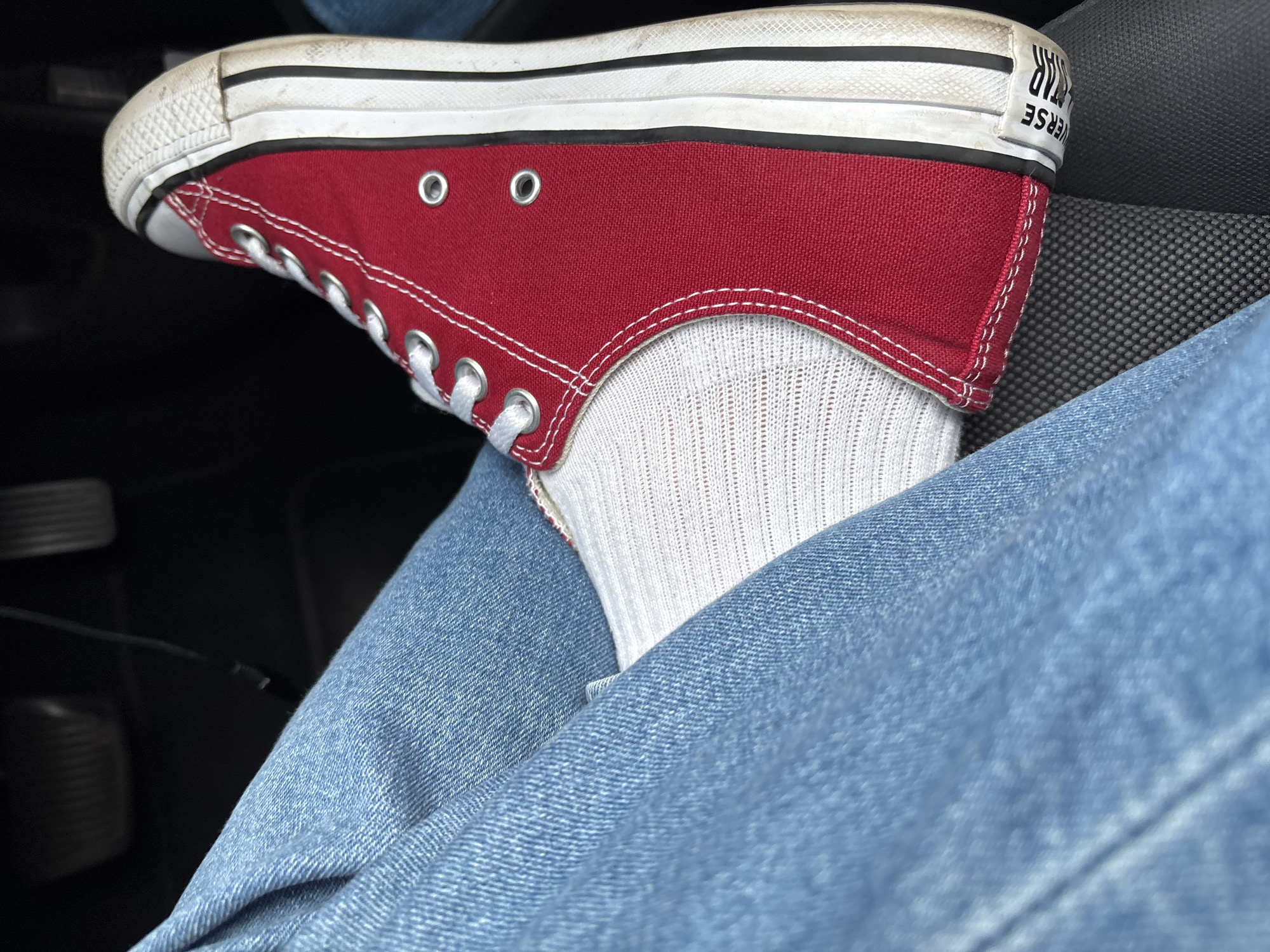 The image shows a close-up of a person's foot wearing a red Converse sneaker and white socks, with blue denim jeans visible. The sneaker has white laces and a white sole with black stripes. The person appears to be sitting, possibly in a car, as indicated by the presence of a dark, textured surface which could be a car seat, and a blurred dark background that might be the car's interior. The lighting suggests an indoor or shaded outdoor environment. There is no visible indication of the specific location within the city of South Staffordshire, United Kingdom.