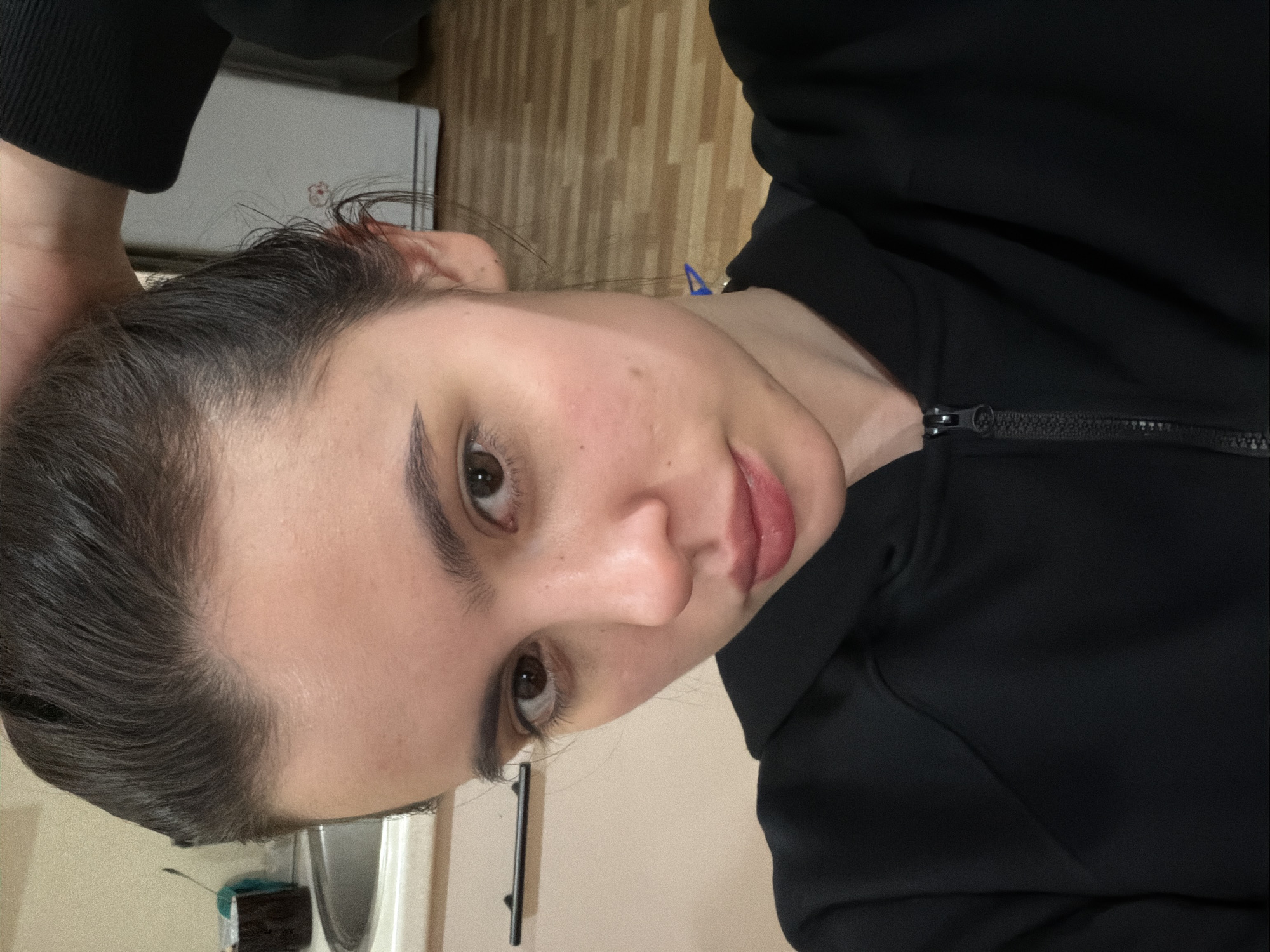 A young woman with dark hair and light skin poses for a selfie in an indoor setting. She is wearing a black zippered jacket. In the background, there is a wooden floor, a sink, and possibly a refrigerator. The location is not discernible as being in the city of Perm, Russia, based on the provided image.