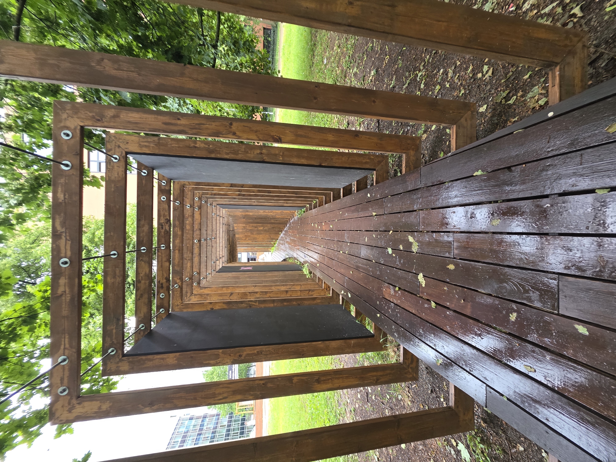 The image displays a long, tunnel-like wooden structure, captured from a slightly elevated perspective looking down its length. The structure is formed by a series of dark brown, square or rectangular wooden frames that progressively decrease in size, creating a strong sense of depth and perspective towards a vanishing point.
The pathway within the structure is made of dark brown wooden planks, which appear wet and somewhat reflective, indicating recent rain or dampness. Scattered green and yellow leaves lie on these wet planks. Along the sides, thin black rods or wires, secured with metal washers and nuts, connect the wooden frames, adding to the architectural detail.
Surrounding the structure is abundant green foliage from trees and green grass on the ground, suggesting an outdoor setting like a park, garden, or campus. Through the gaps in the wooden frames and the trees, modern buildings with visible windows can be seen in the background, indicating an urban or suburban environment.
The scene is set during daytime, with diffused light. No individuals or discernible activities are present. The atmosphere is quiet and natural, showcasing an interesting architectural feature integrated into an outdoor space. There is no visible text.