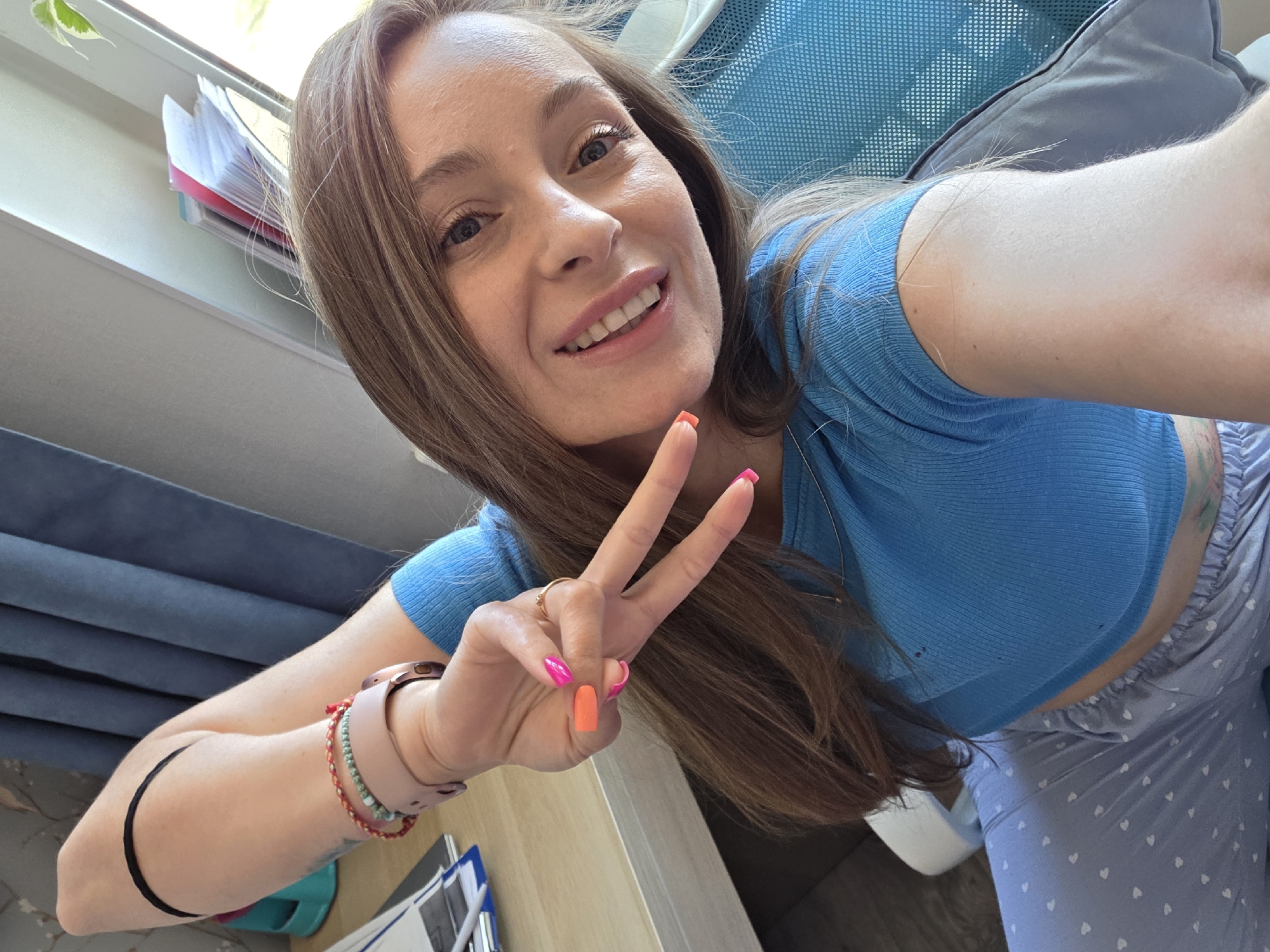 A woman is taking a selfie, giving a peace sign. She has long brown hair and is wearing a blue top and patterned pants. She has painted her nails in different shades of pink and orange. She is wearing a watch and bracelets on her left wrist and a ring on her left ring finger. The background shows part of a window with papers, a blue curtain, and a blue chair. The location and city cannot be determined from the image.