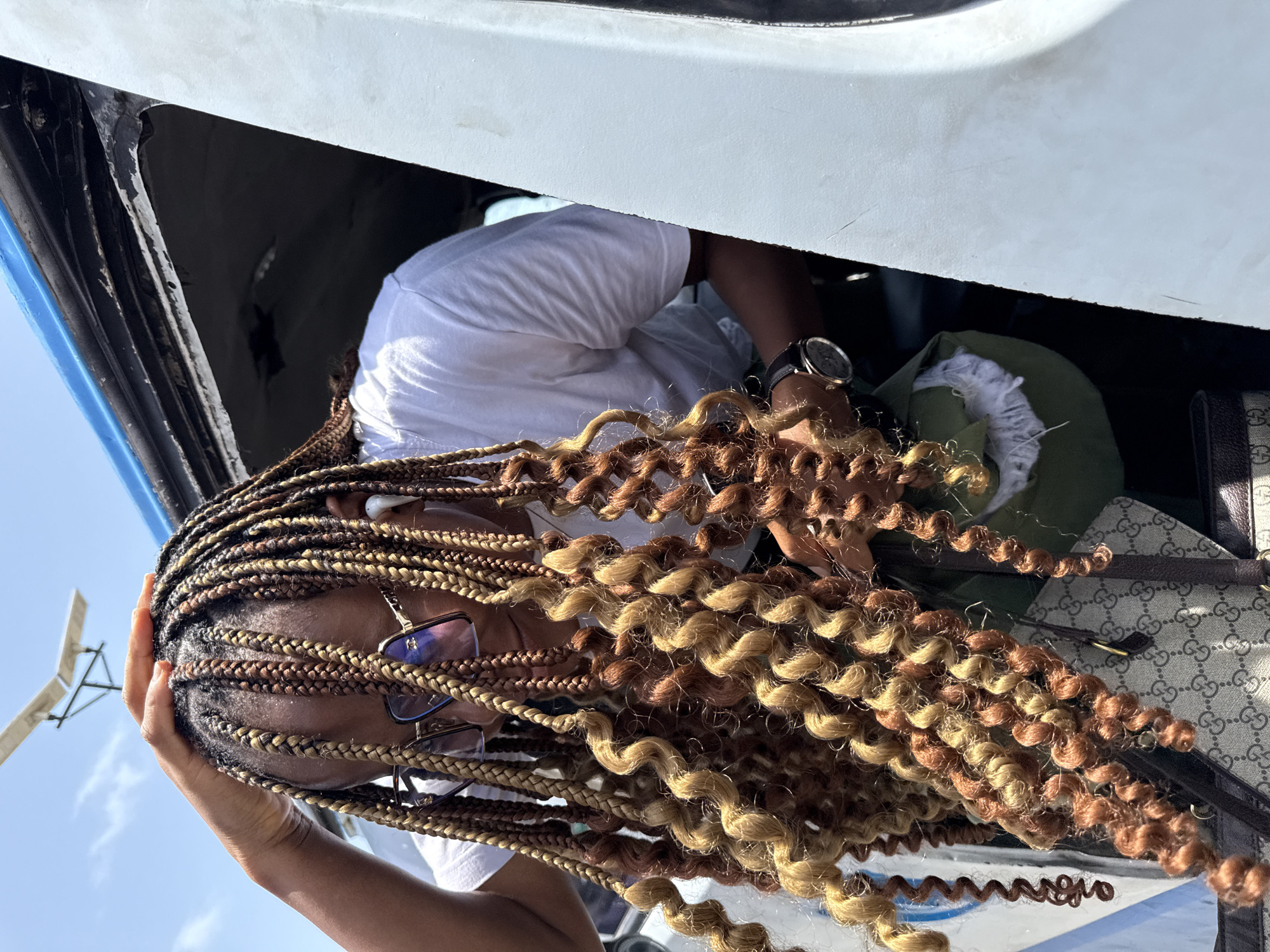 A person with long, curly, two-toned brown and blonde braids is visible, wearing glasses and a white shirt, looking into a vehicle in Ilado, Nigeria. A black watch is on the person's wrist, and a Gucci bag is partially visible in the background, along with an olive green bag. The sky is visible in the background.